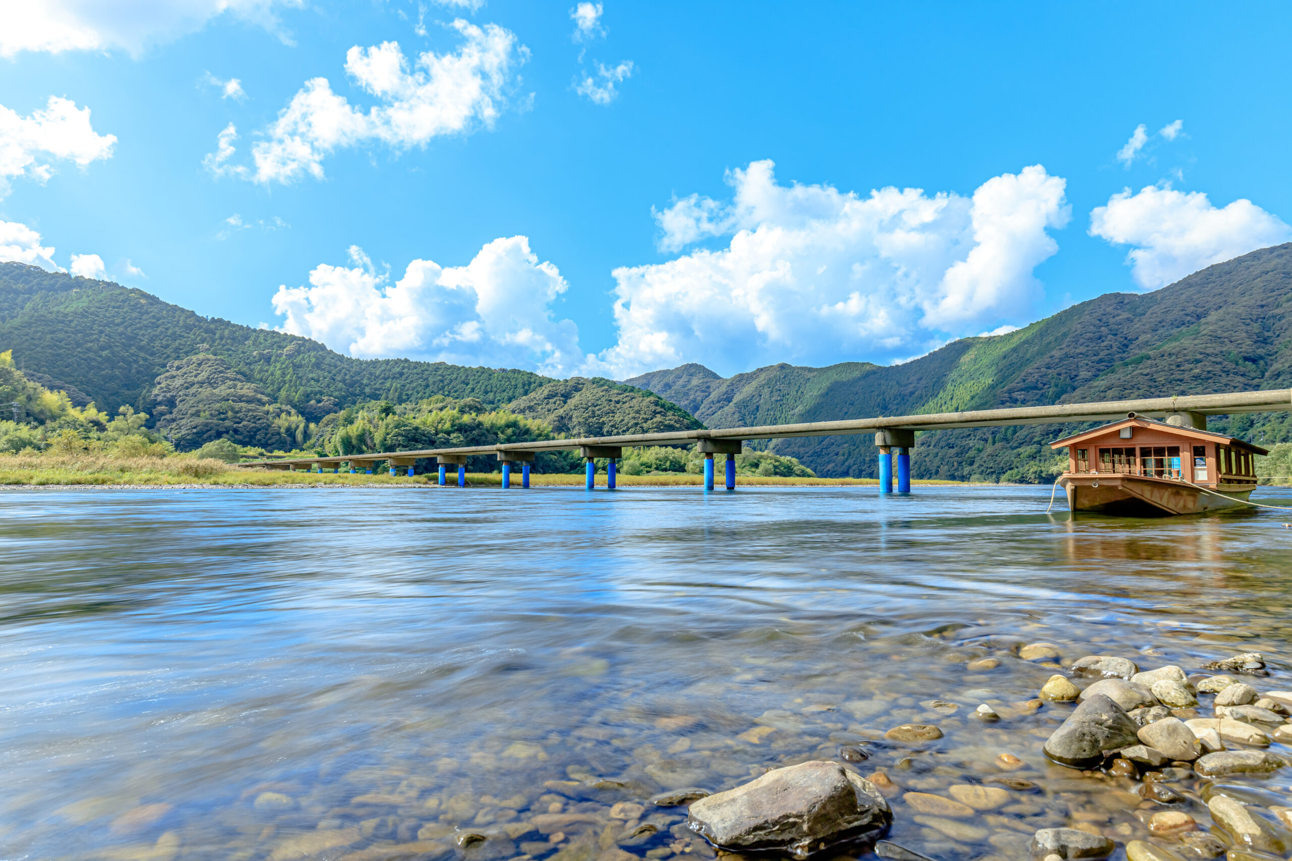 Wide river scene with a long bridge on blue piers spanning the water, and a small wooden building on the right, set against green mountains and a bright sky.