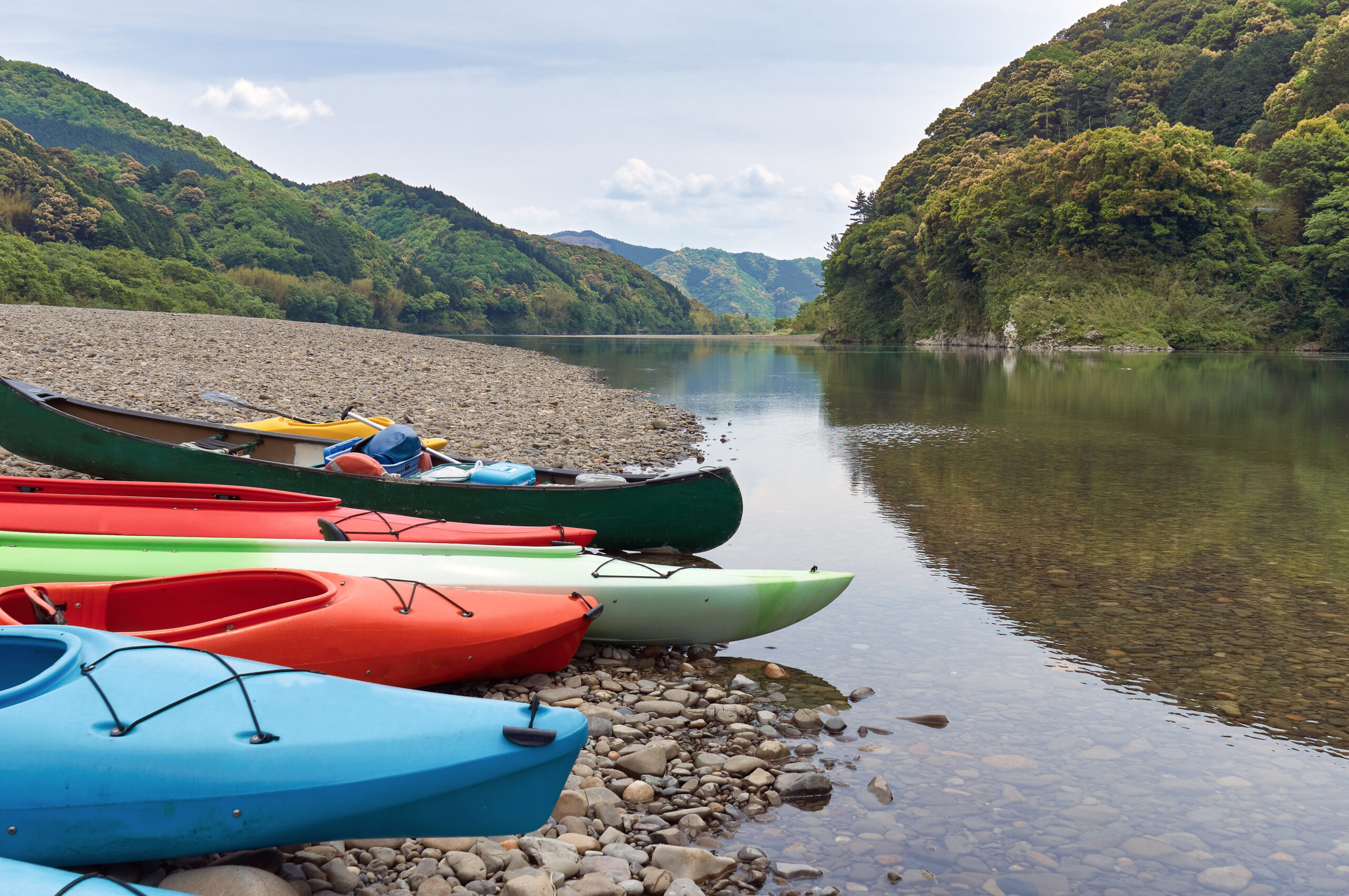 Colorful kayaks lined up on a pebbled shore beside a calm river with green forested hills in the background.