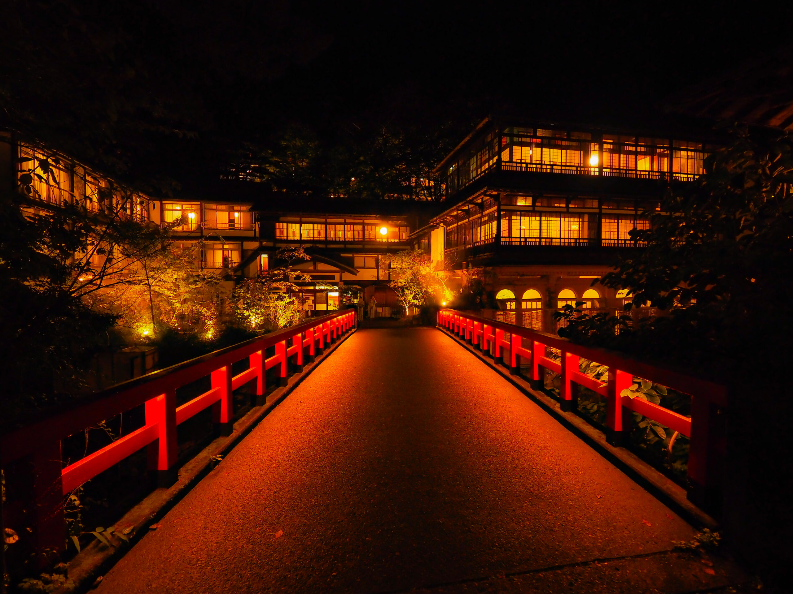 Nighttime view of a lit, multi-story wooden building with warm orange lights, seen from a red railing bridge walkway.