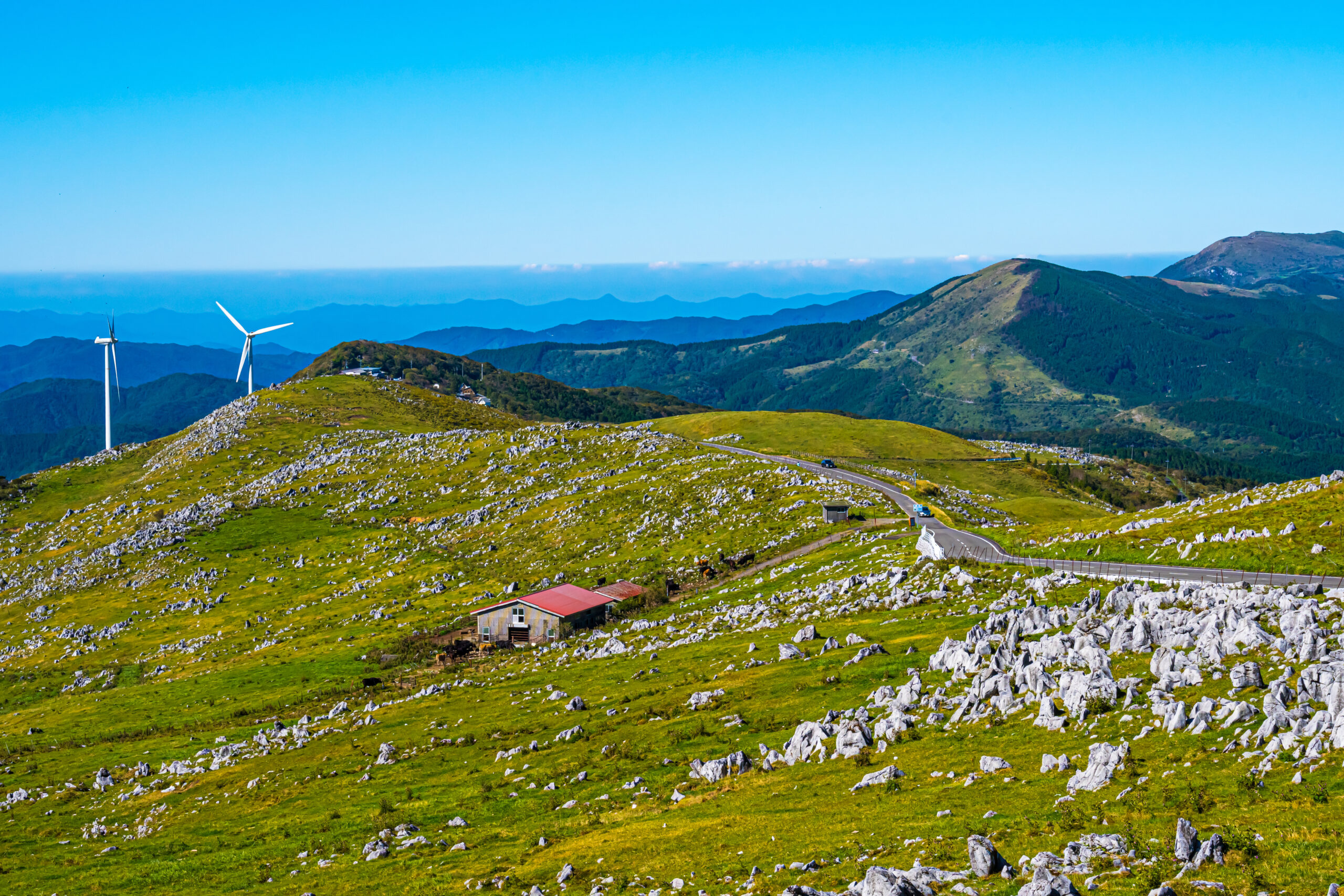 Wind turbines on a grassy, rocky hillside with a winding road and distant blue mountains under a clear sky
