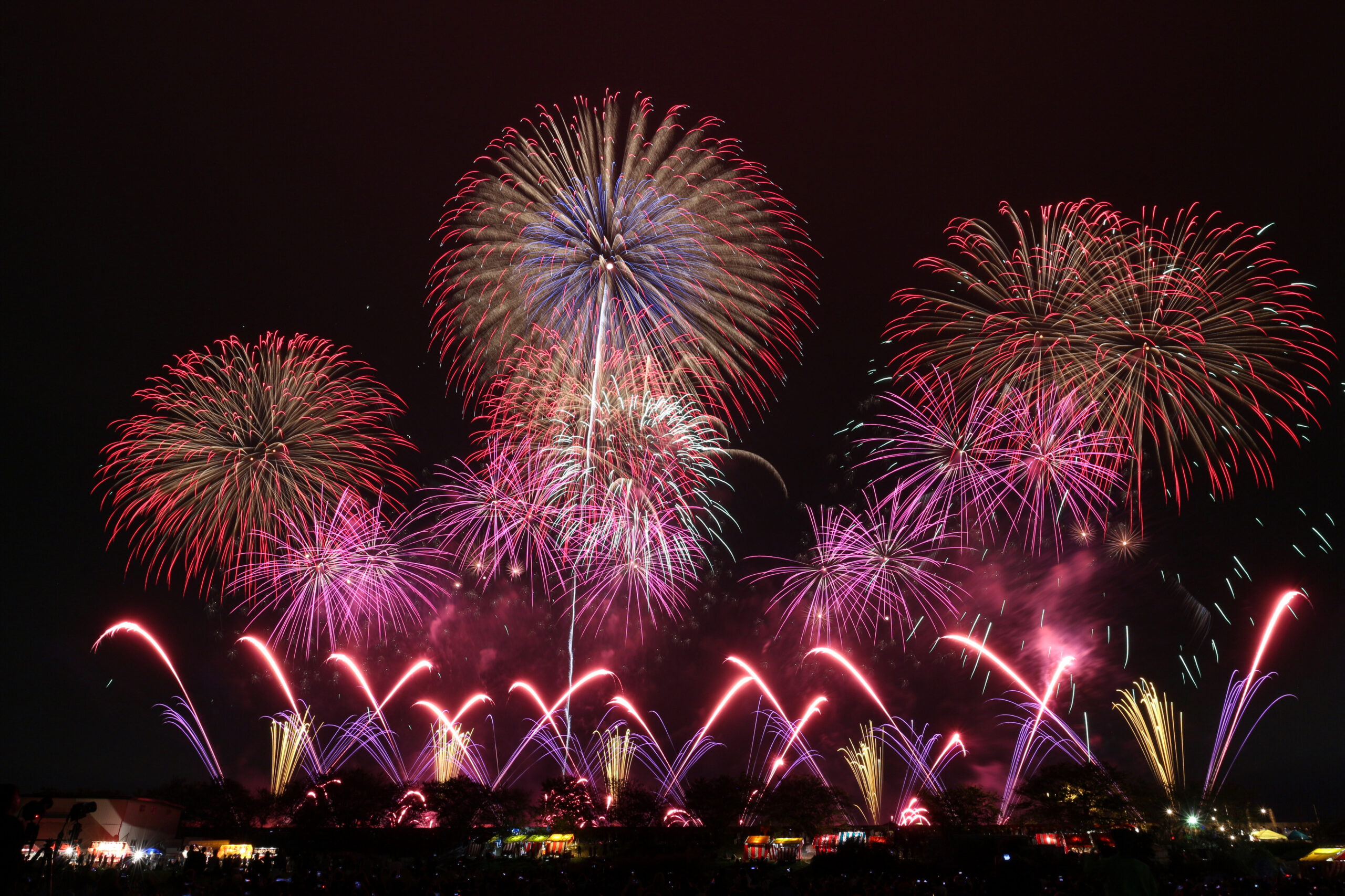 Nighttime fireworks show over a crowd with colorful bursts lighting the sky, red, blue, and purple fountains soaring above festival tents.