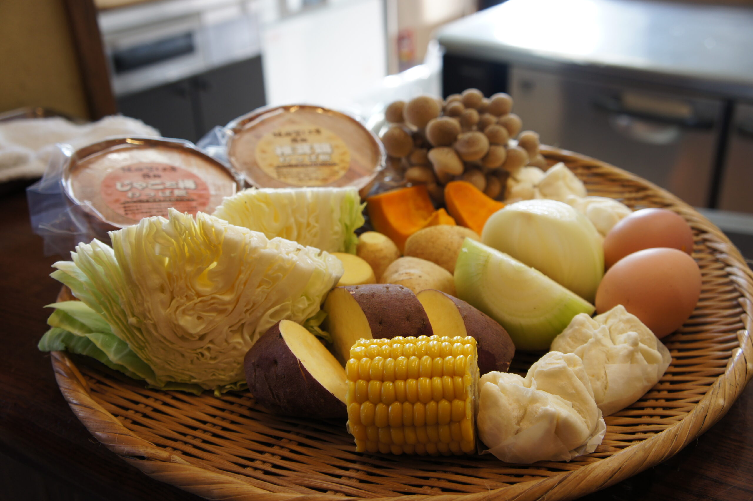 Basket of fresh groceries and cheeses on a countertop, featuring corn on the cob, cabbage, potatoes, onions, and small round cheeses or mushrooms clustered in the background.