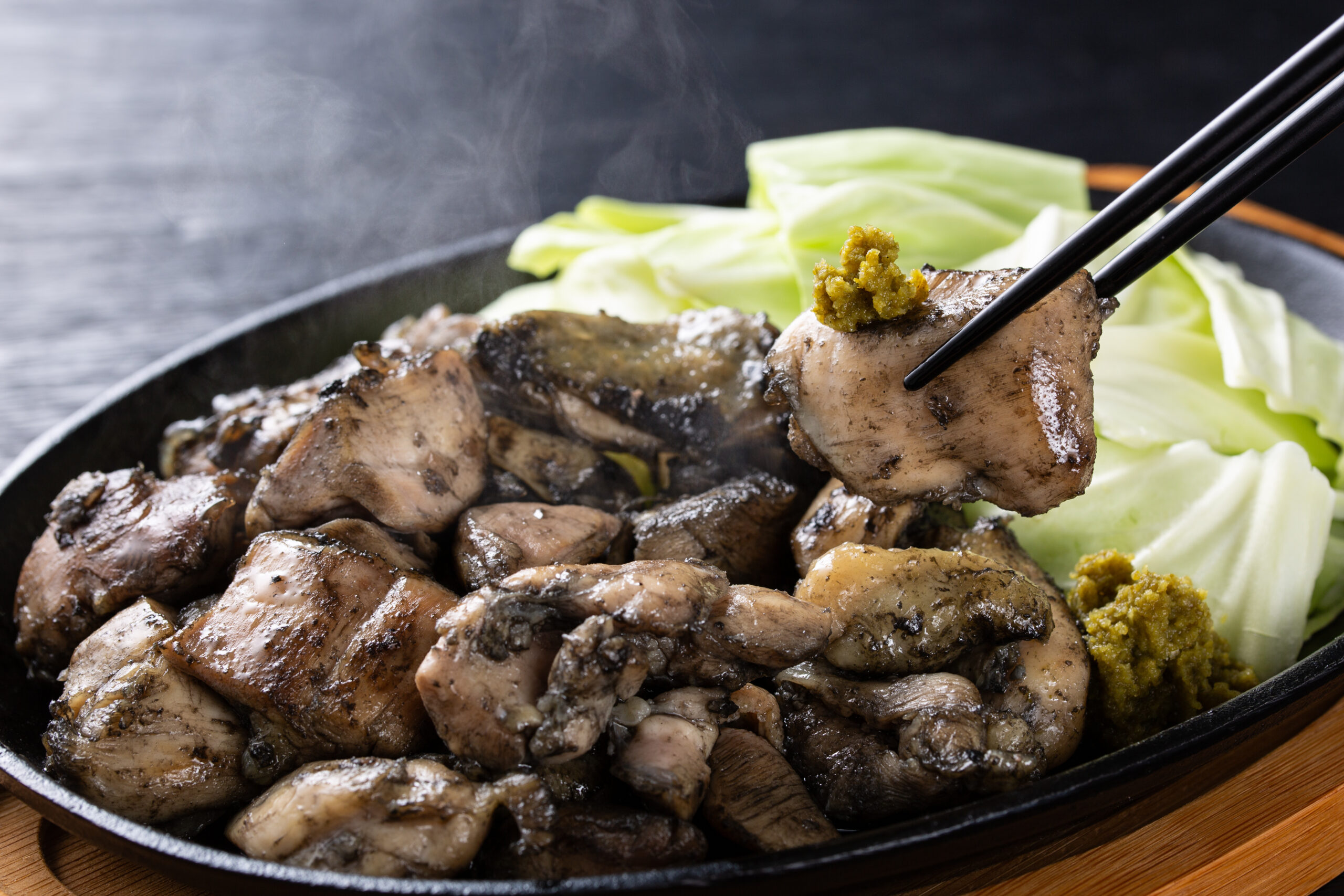 Sizzling plate of grilled meat chunks with pale cabbage leaves in the background; chopsticks lifting a piece with a dollop of mustard.