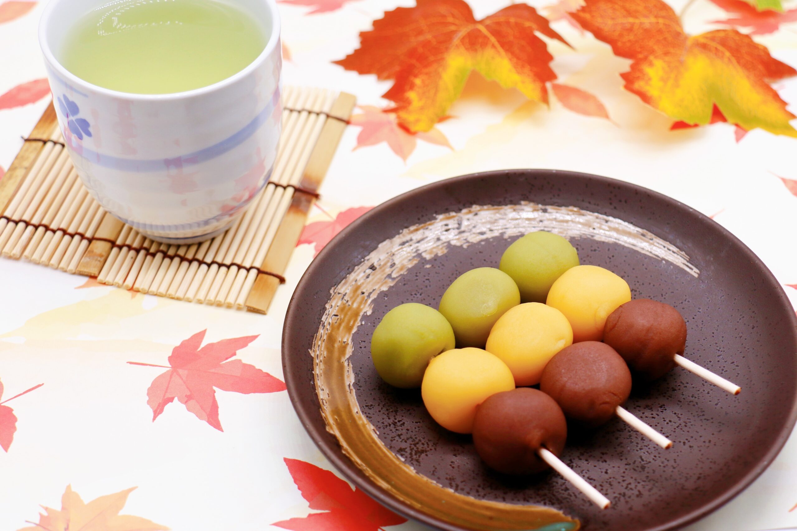 Skewered dango mochi in green, yellow and brown on a brown plate beside a cup of green tea and autumn leaves in the background.