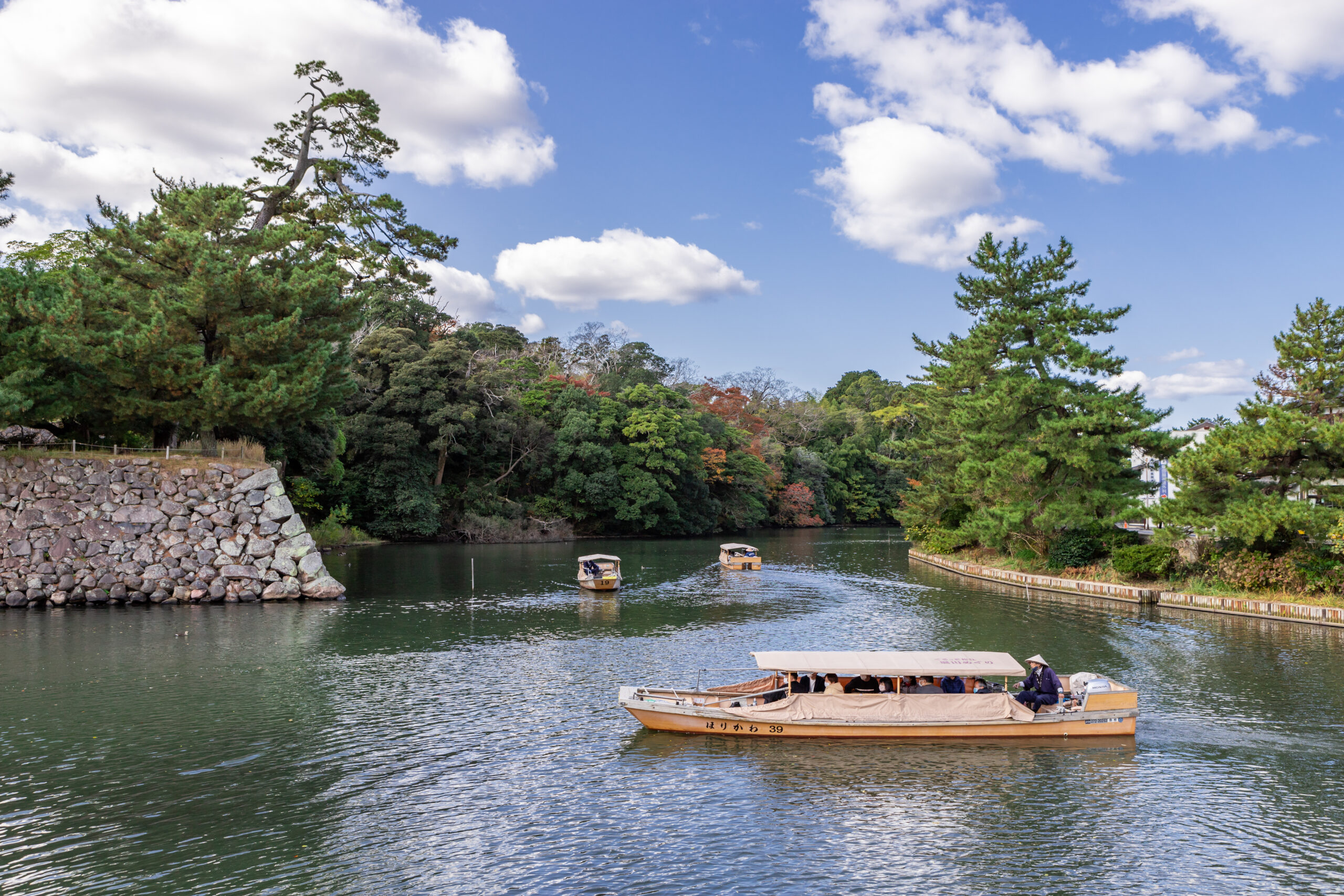 Passenger boats with beige canopies glide on a calm lake, edged by a rocky wall and dense greenery under a blue, partly cloudy sky.