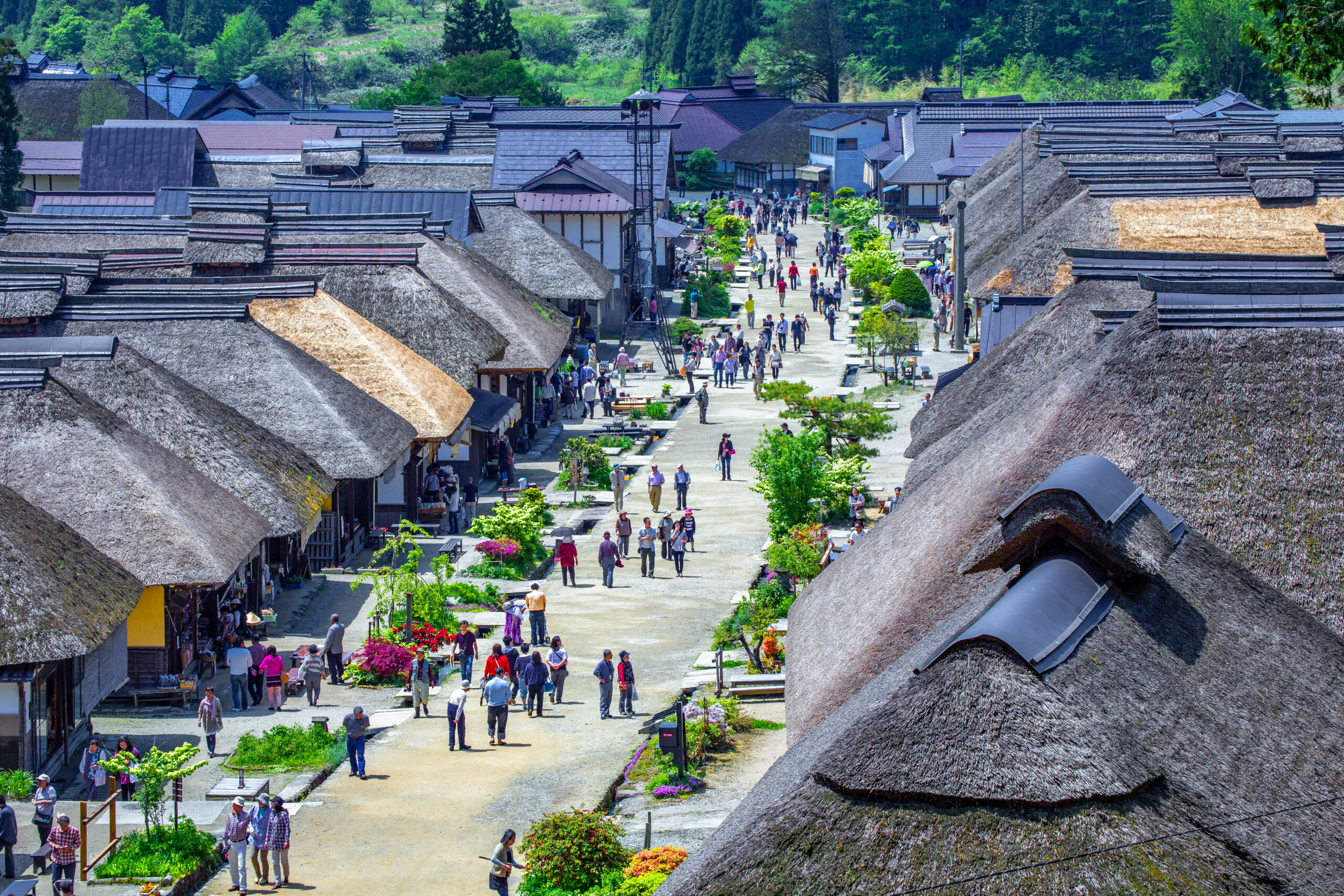 Wide street lined with thatched-roof houses and greenery; many people stroll and shop along the path.