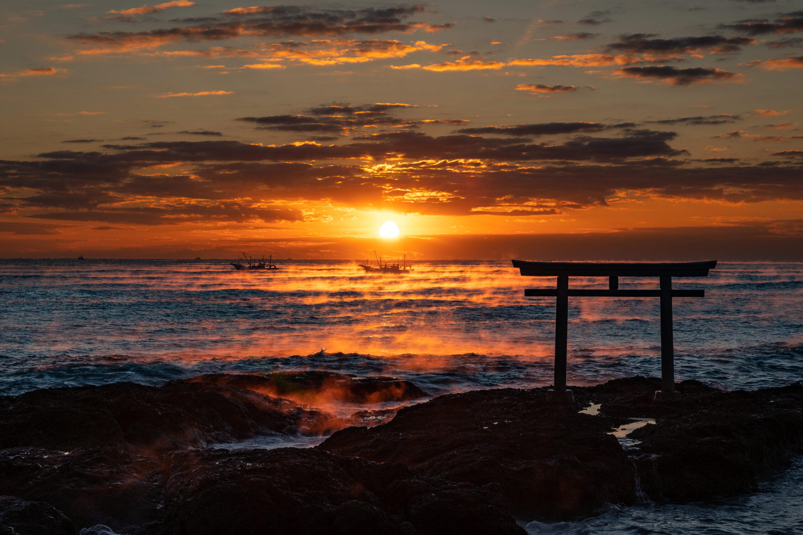 Fiery sunset over the ocean with a rocky foreground and a wooden gate structure in the foreground, boats on the horizon.
