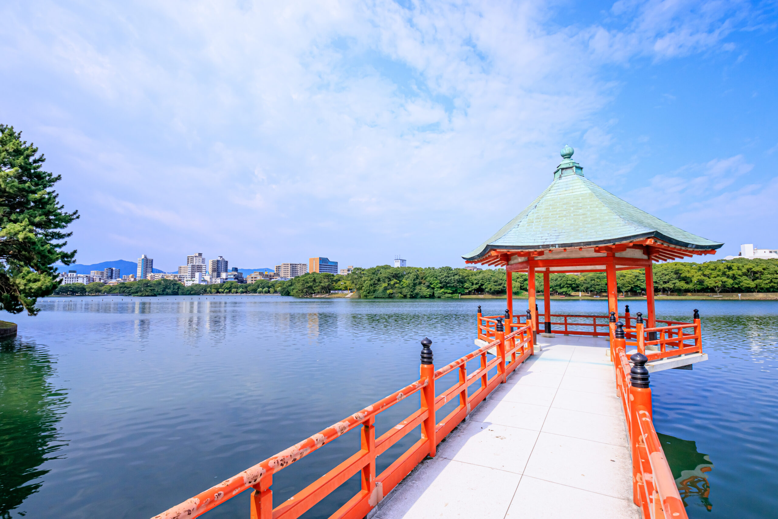 Red pavilion on a pier extending over a calm lake with a city skyline in the distance under a blue sky.