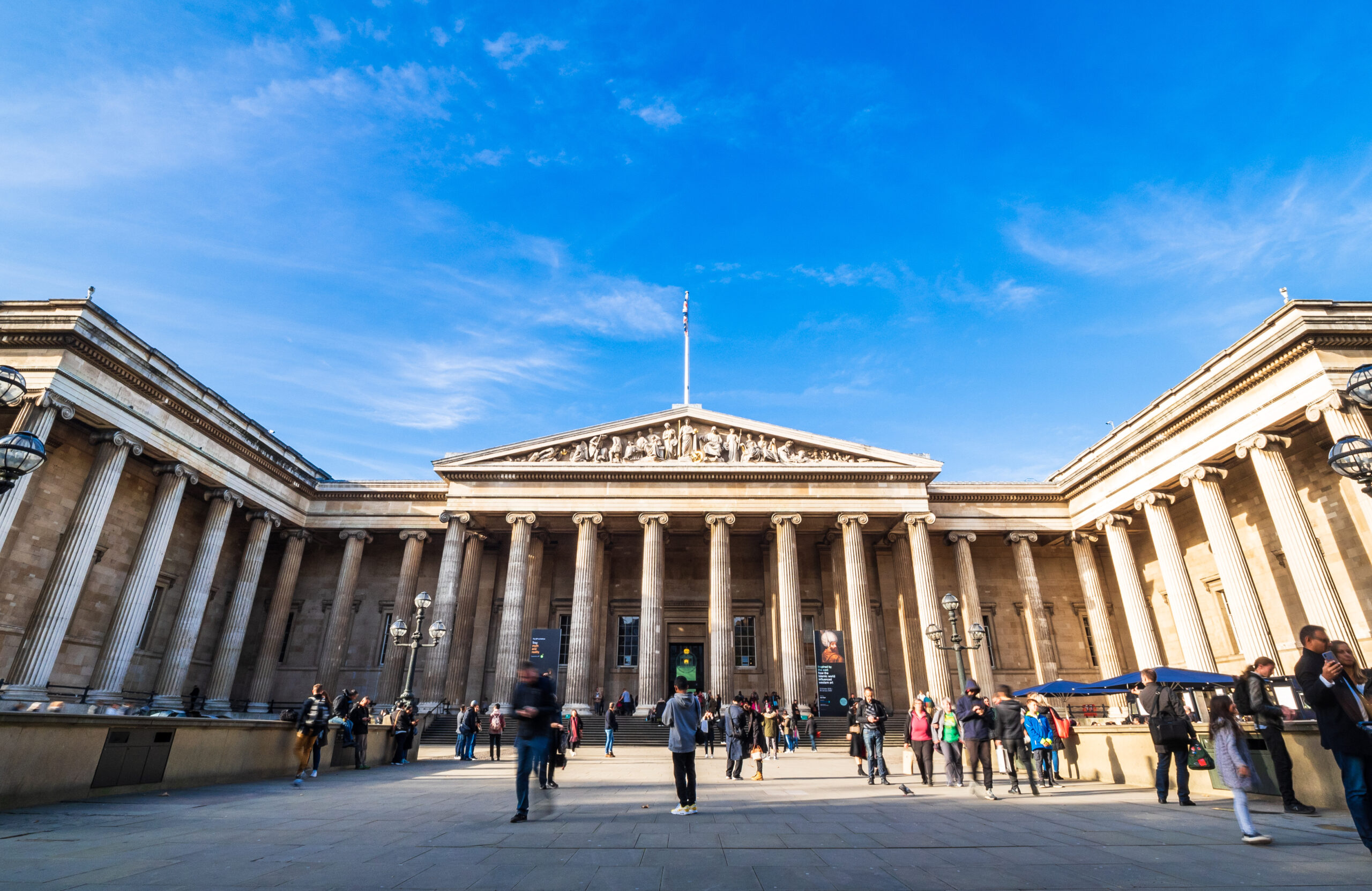 Front view of a grand neoclassical building with a columned portico, sculpted pediment, and a flag atop; blue sky above.