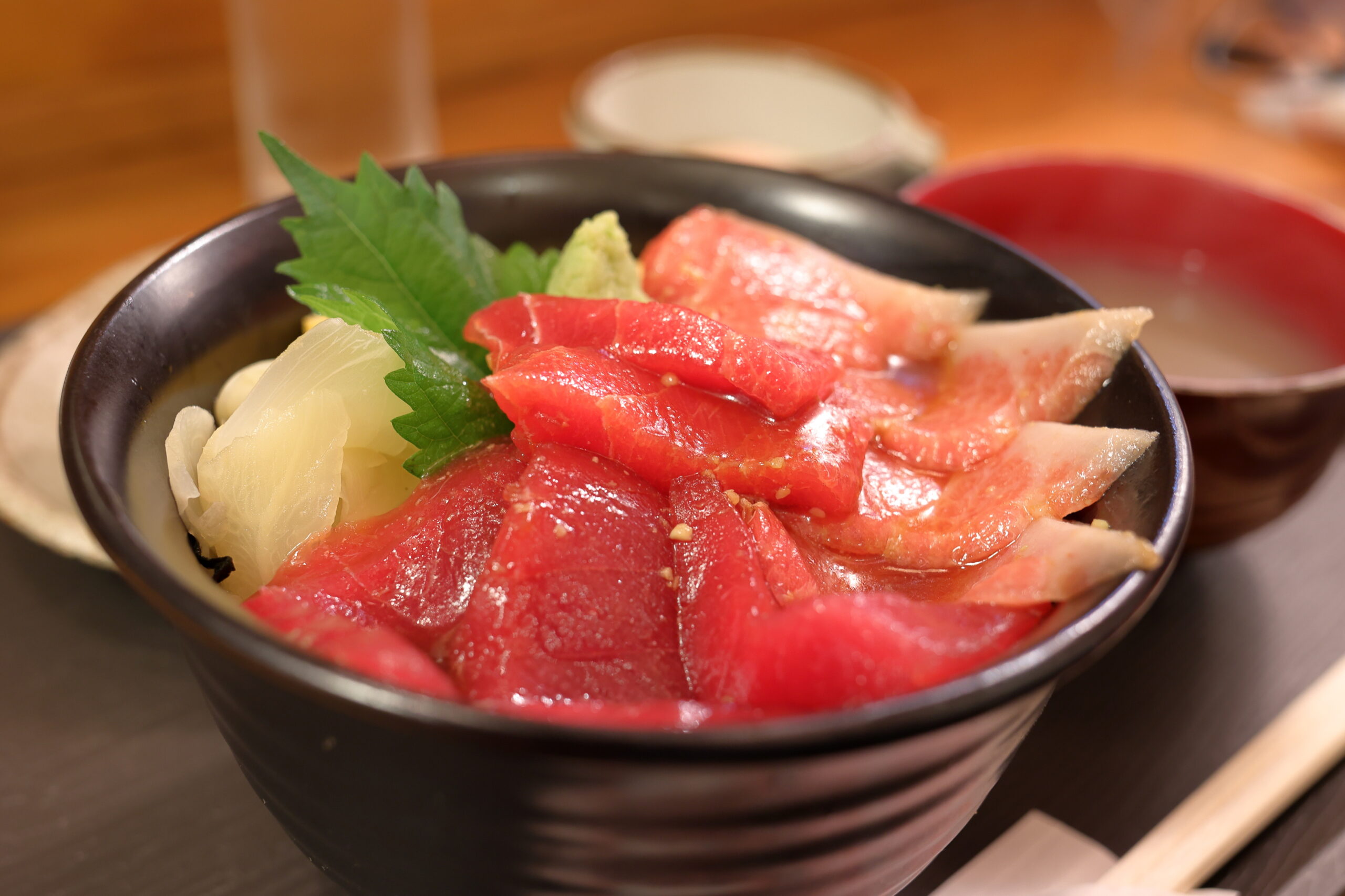 Bowl of tuna sashimi with wasabi, white onion, and shiso leaf in a black bowl.