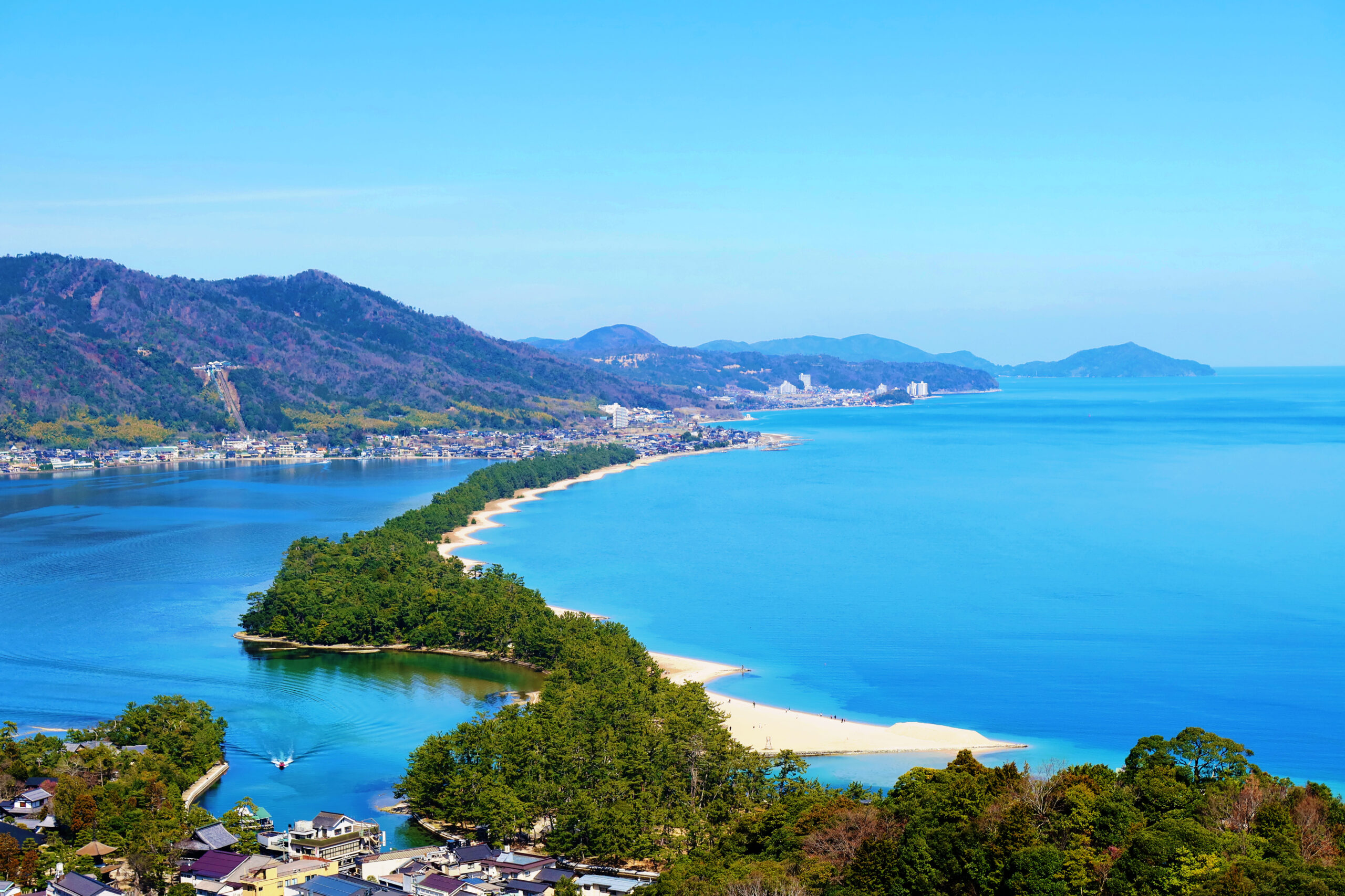 Aerial view of a coastal town along a curved sandy beach, with forested hills and a calm blue sea under a clear sky.