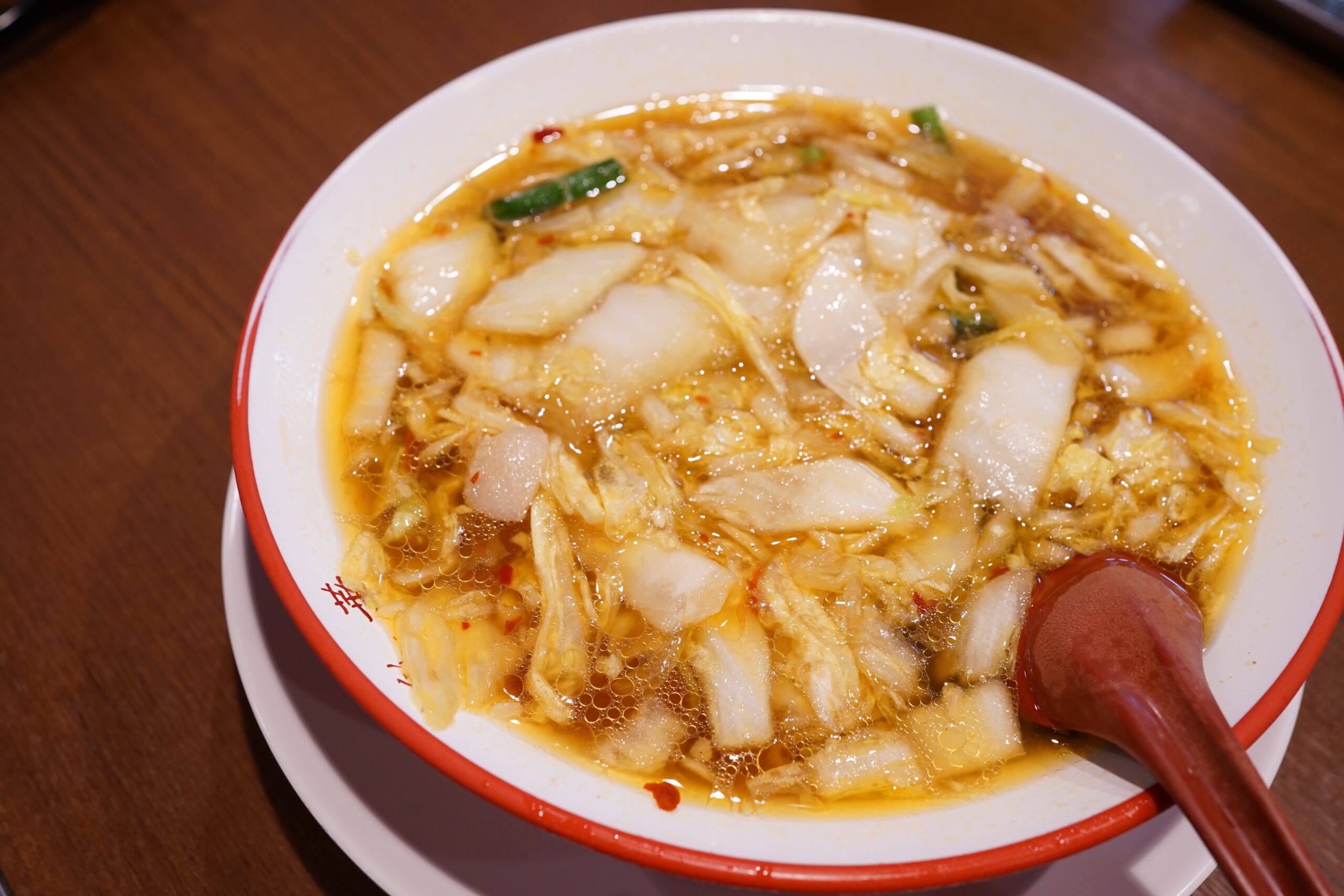 Bowl of spicy Sichuan-style soup with cabbage, vegetables, and chili oil in a red-edged white bowl, ladle resting inside.