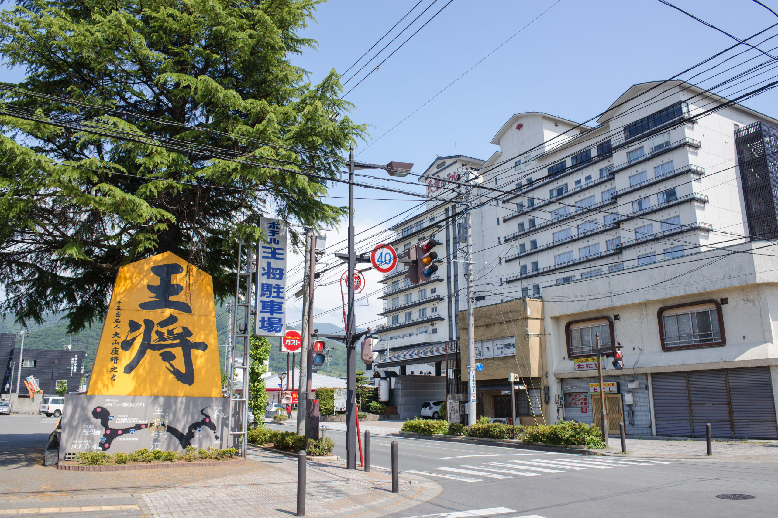 Urban street scene in Japan with a large green tree, overhead power lines, and a white multi-story building in the background, and a tall yellow sign with black kanji in the foreground.