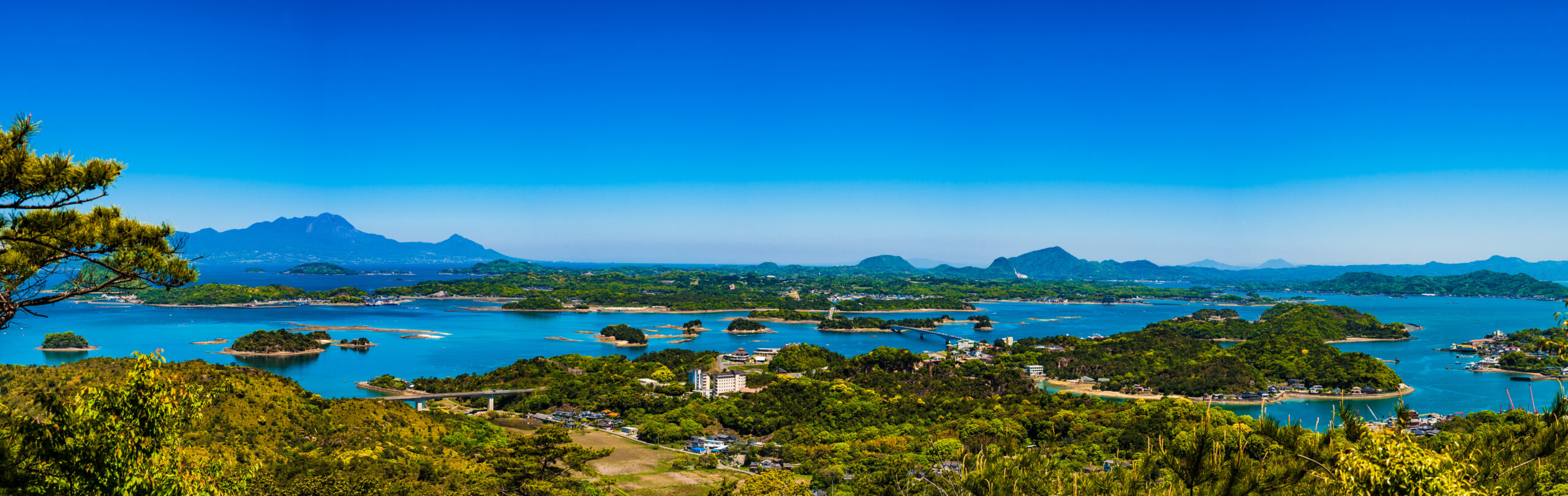 Panoramic view of a blue sea with many small green islands and distant mountains under a clear sky.