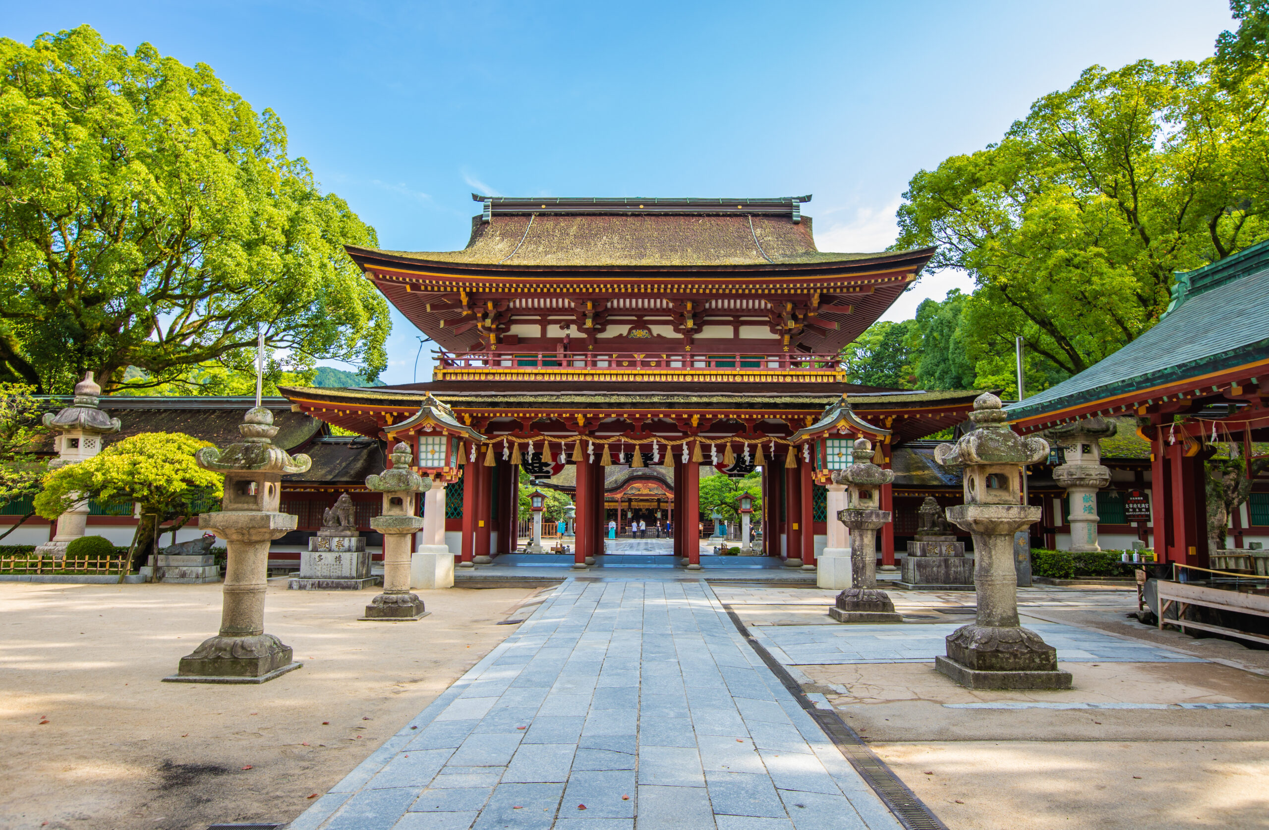 Temple shrine entrance with red wooden gates, stone lanterns, and lush green trees under a bright blue sky.