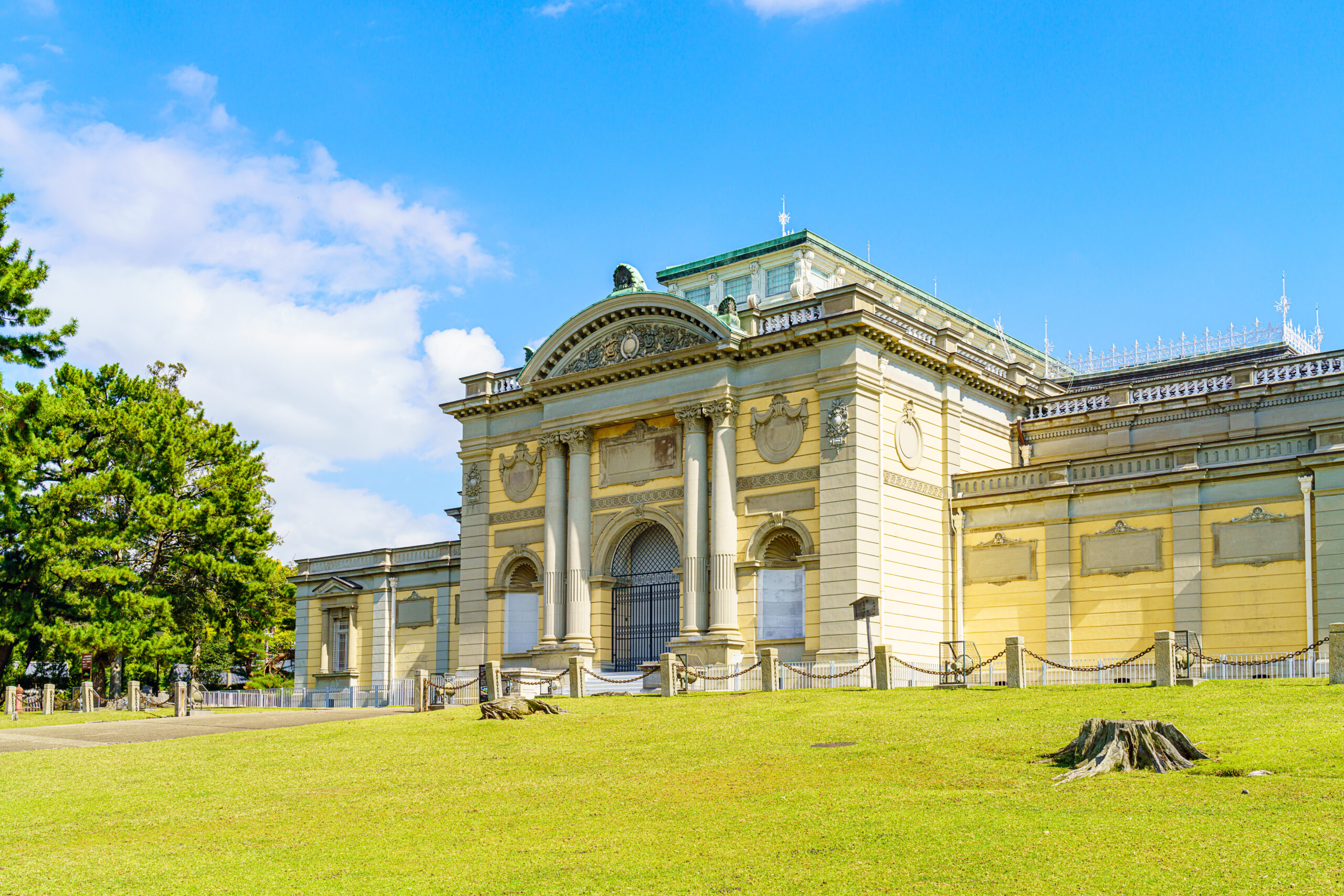 Historic neoclassical building with arched entrances and ornate stonework, set on a green lawn under a bright blue sky.