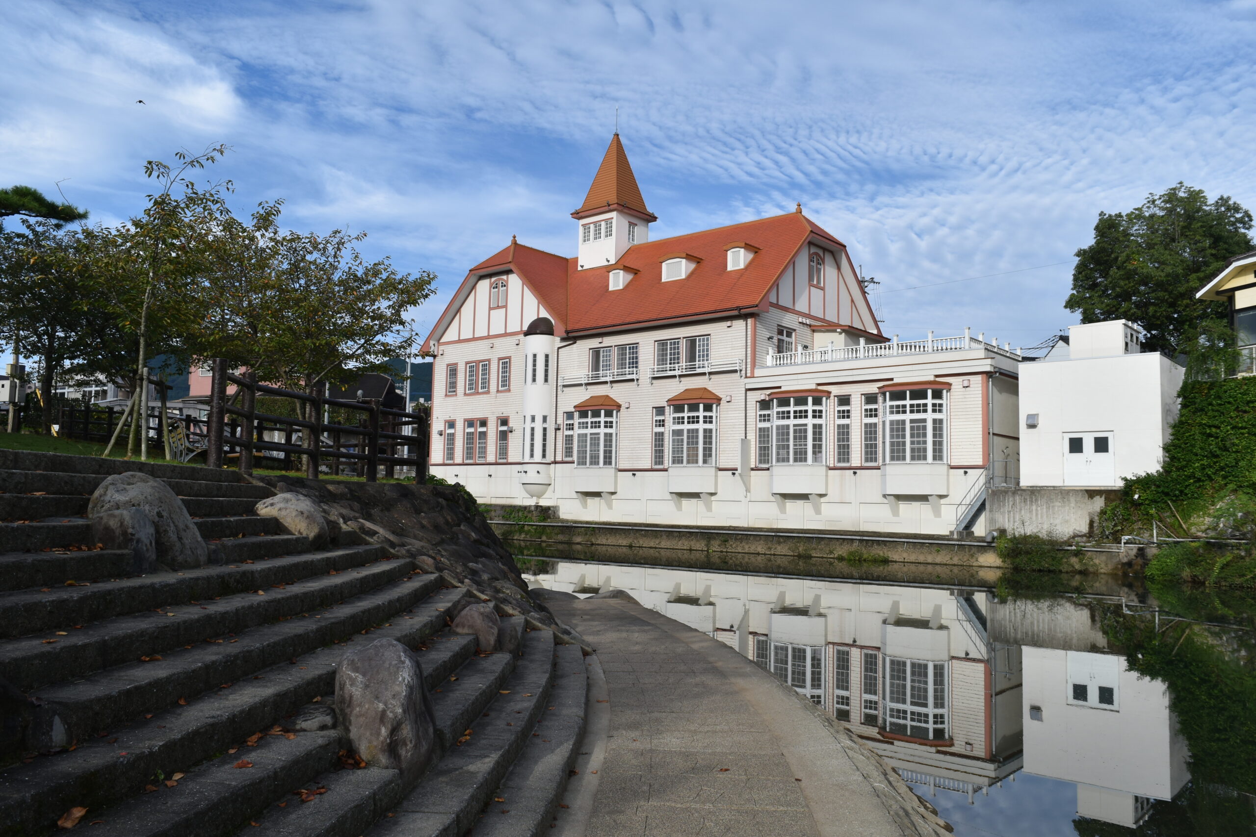 White building with red roof and tower beside a calm waterway, reflections visible in the water.