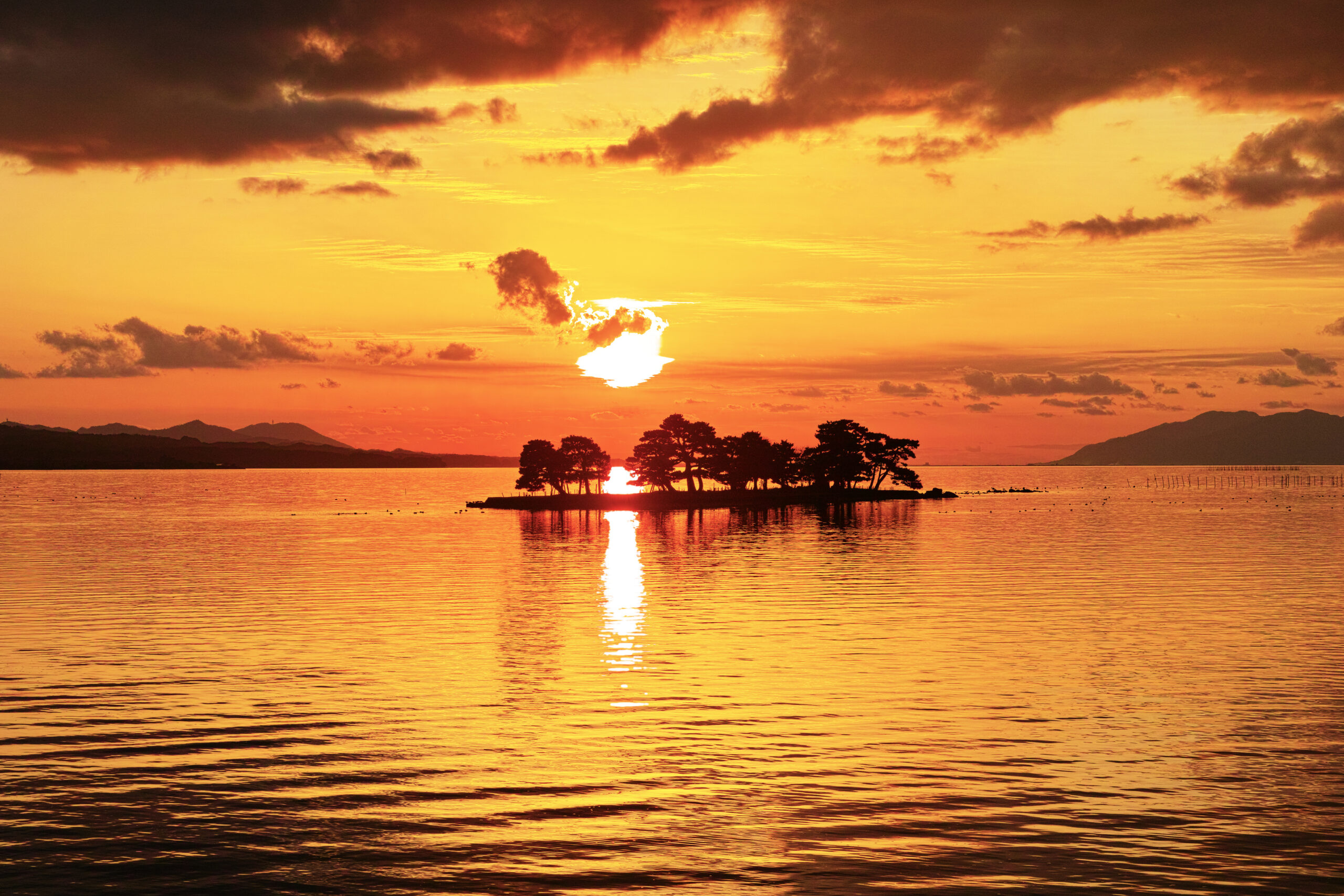 Sunset over a calm body of water with a small, tree-covered islet silhouetted against an orange sky and its reflection on the water
