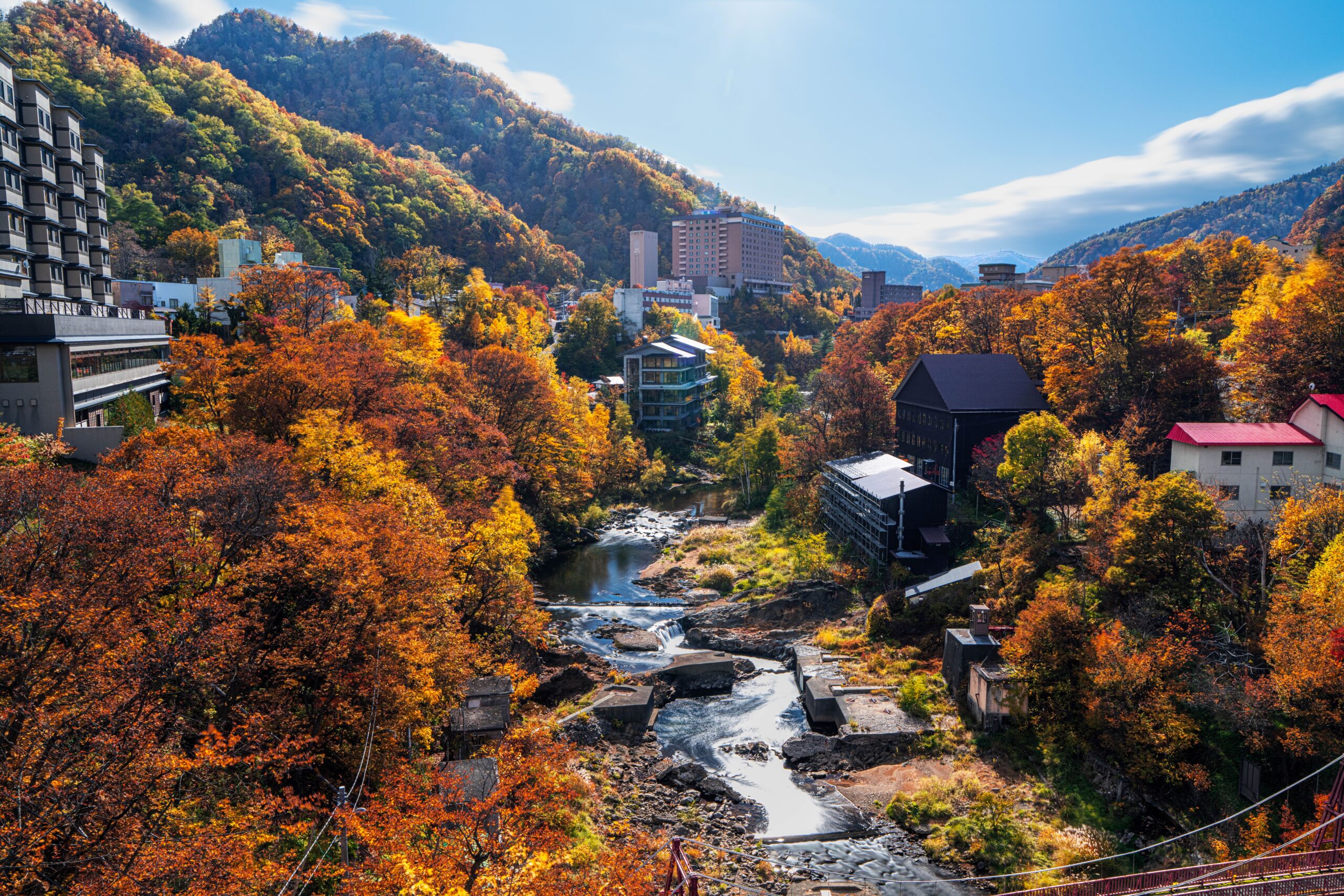 Panoramic view of a valley town with colorful autumn trees, a winding river, and mountains in the background.",