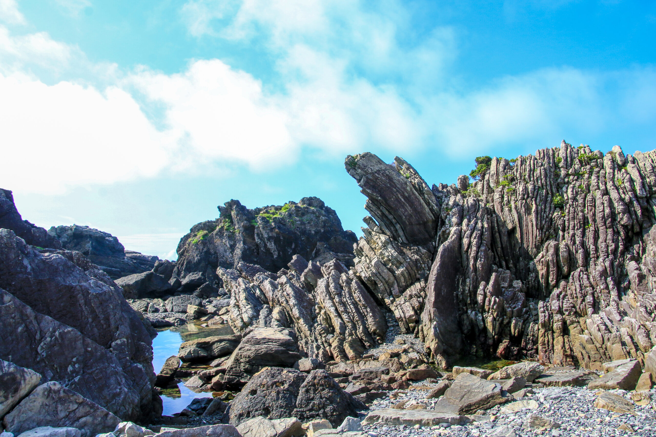 Jagged basalt rock formations along a rocky shoreline under a blue sky.