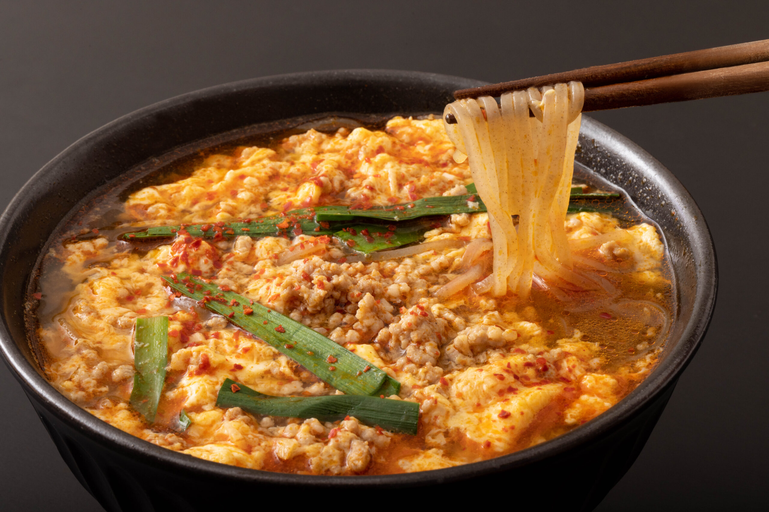 Clay pot filled with spicy mapo tofu and minced meat, garnished with green onions, with noodles being lifted by chopsticks