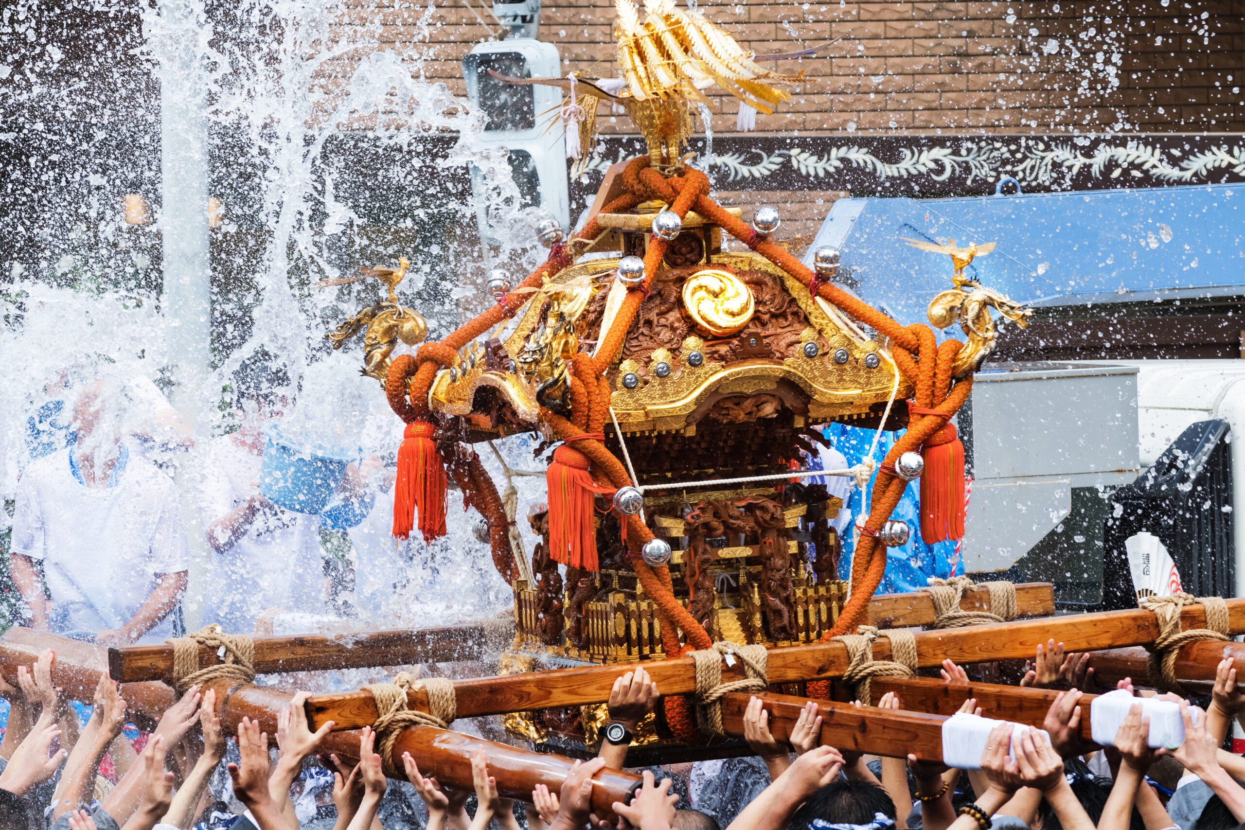Ornate golden festival float with red rope decorations being carried through a crowded scene as water splashes around it.