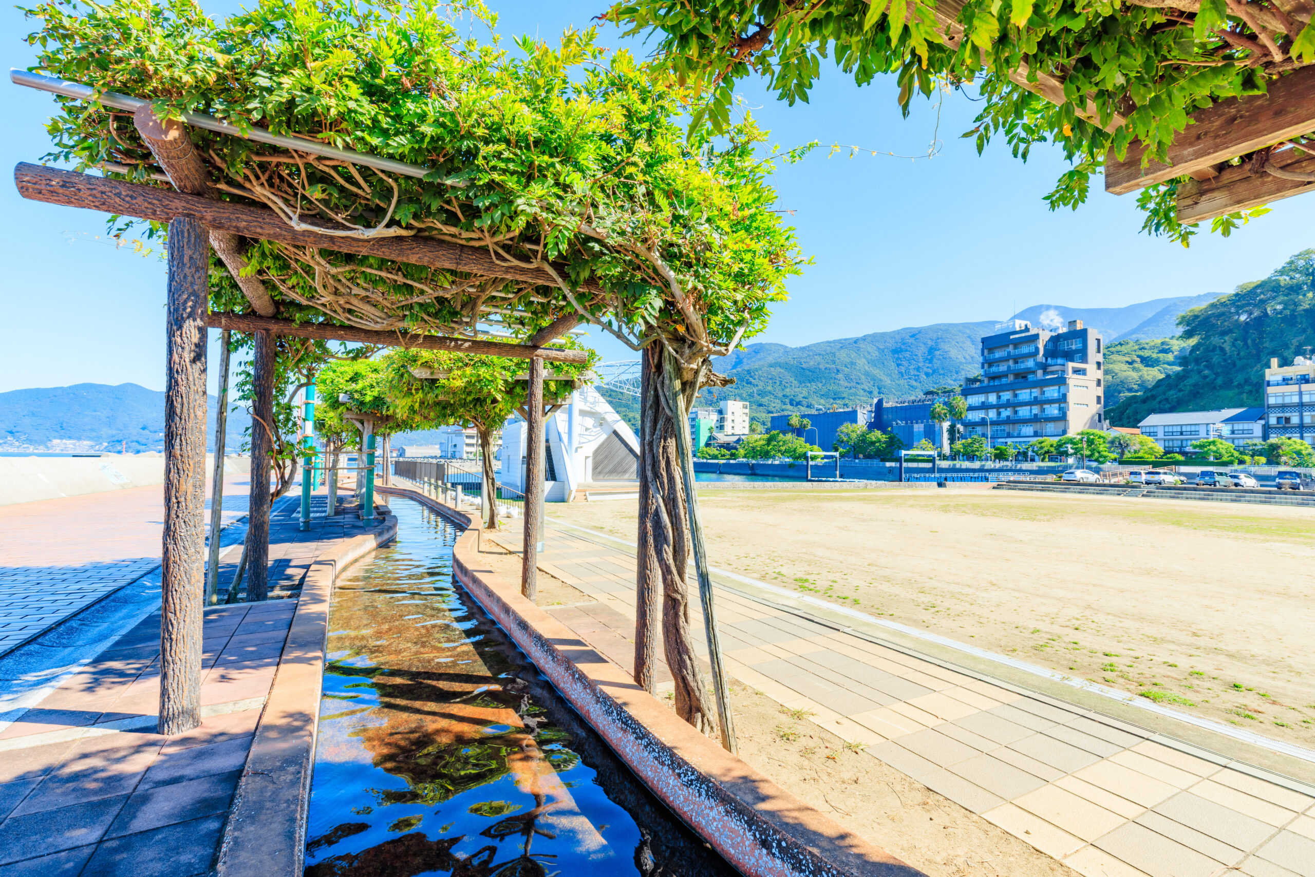 Tree-covered walkway with a narrow reflecting water channel beside a tiled path along a city waterfront.