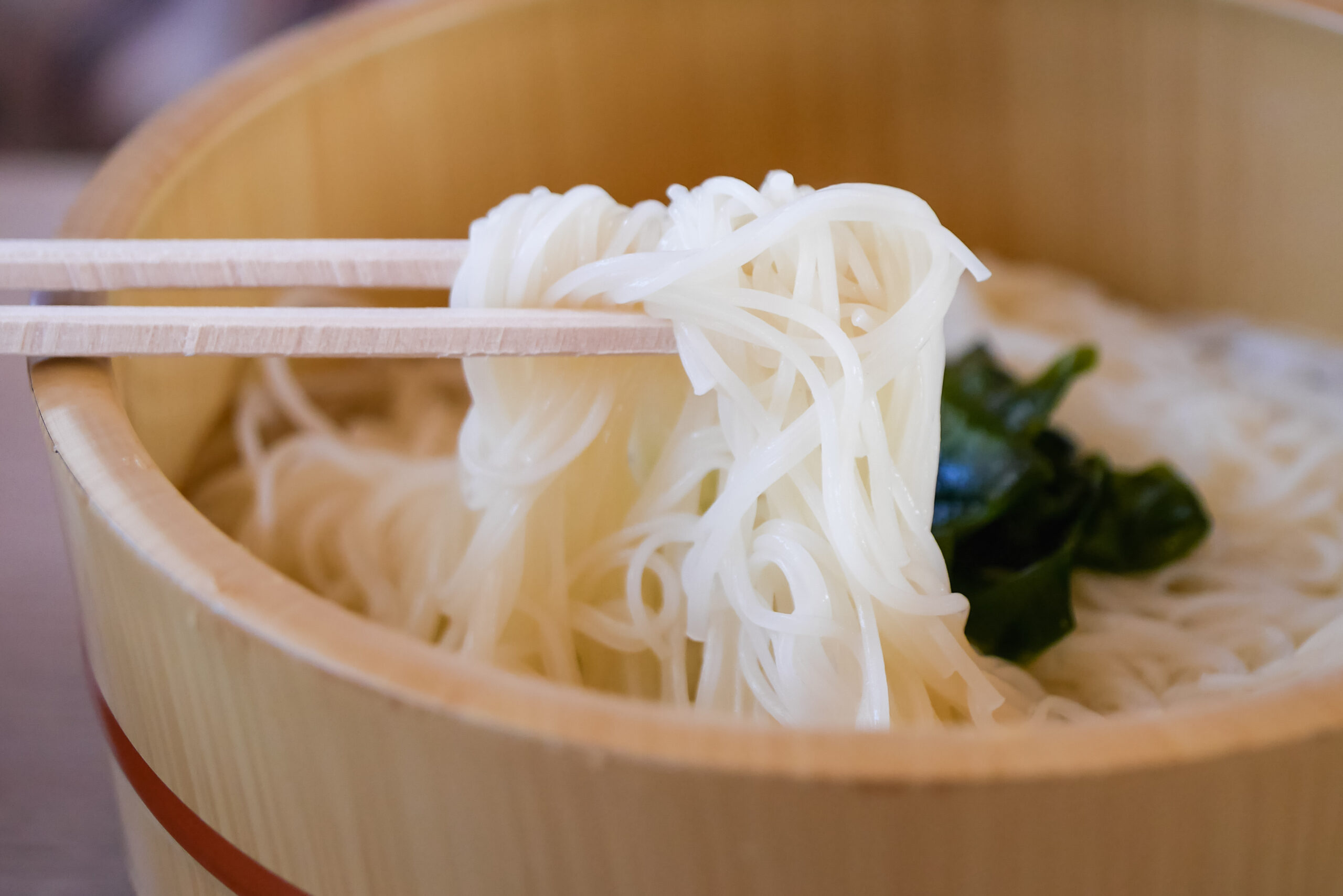Chopsticks lift a strand of white noodles from a wooden bowl.