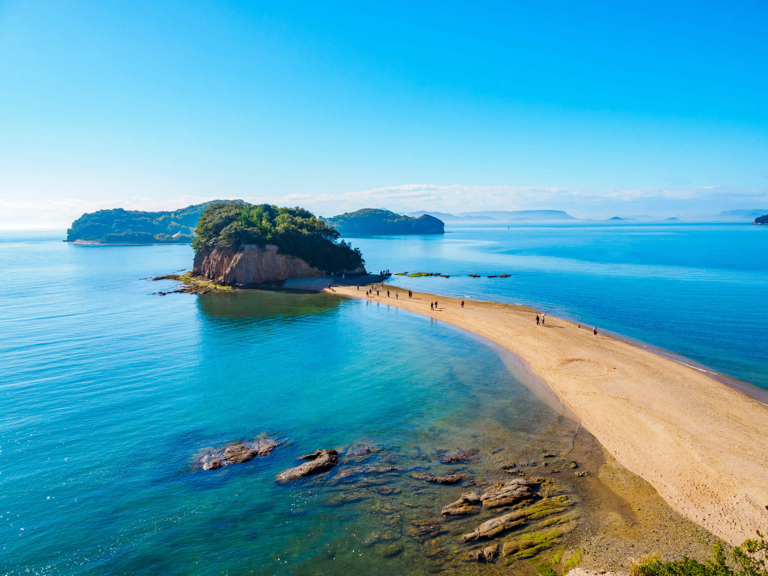 Clear blue sea with a sandy spit and a small, tree-covered island jutting from the water under a bright sky.