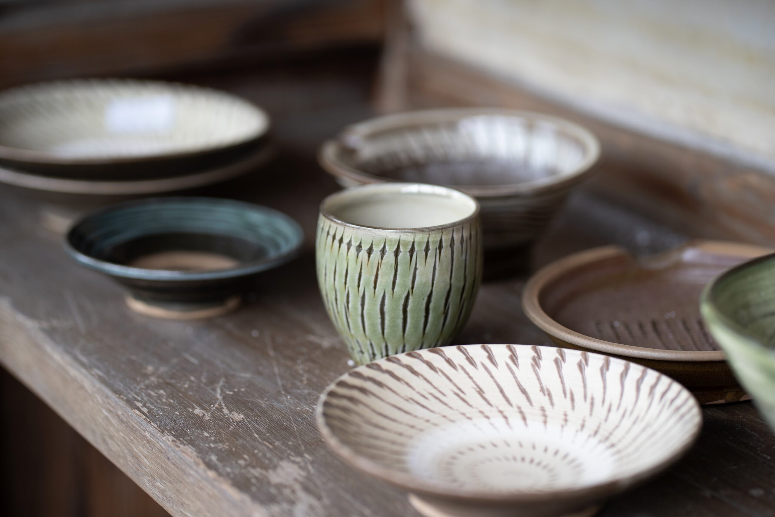 Stacked ceramic bowls and plates on a rustic wooden table, photographer’s focus on a green striped cup in the foreground.