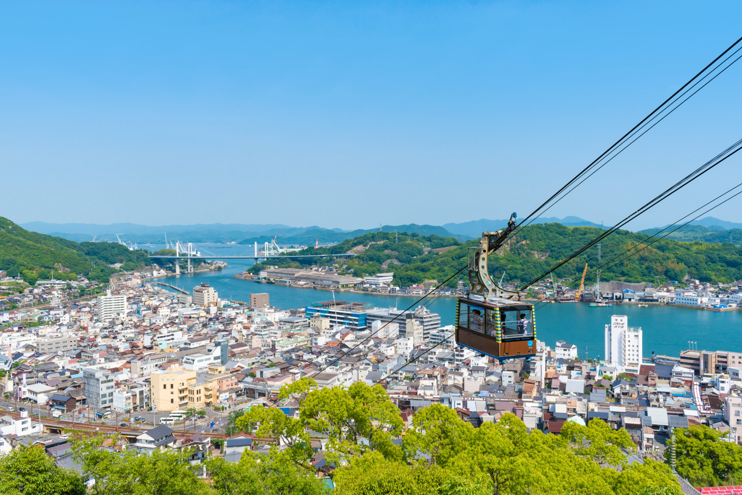 Cable car gondola suspended over a densely built city with a river, bridges, and green hills under a blue sky.