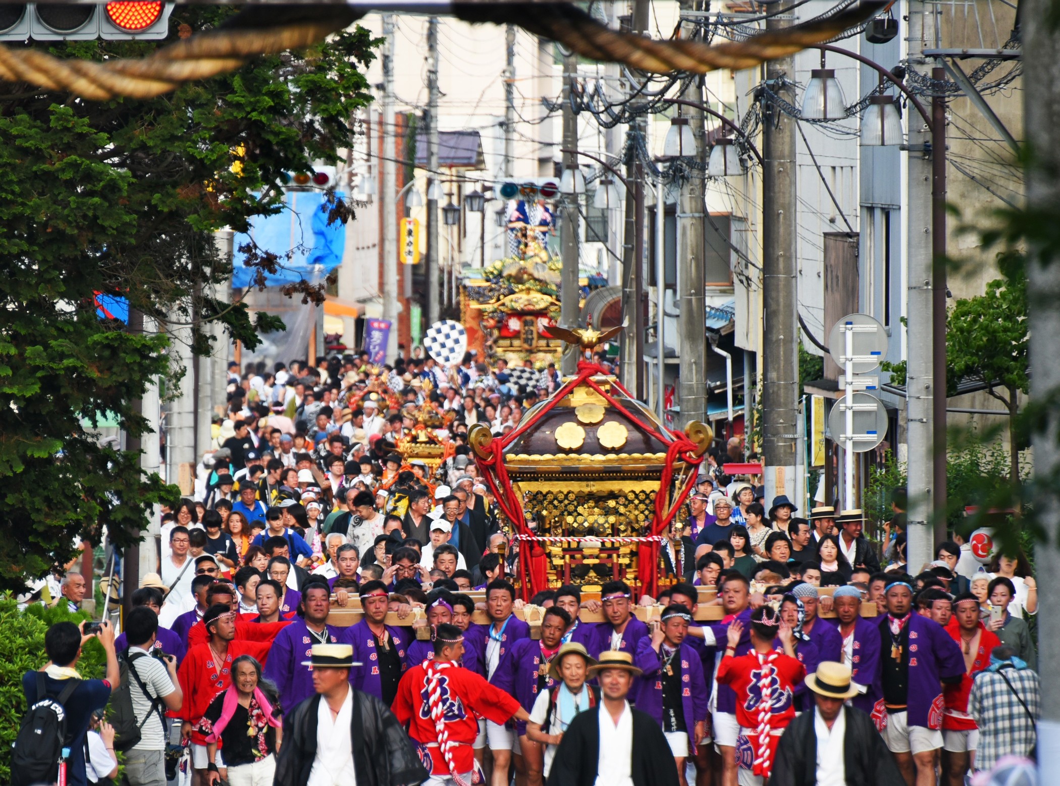 Crowd of people in colorful traditional clothing marching down a crowded urban street during a festival, with a decorative float visible in the center.