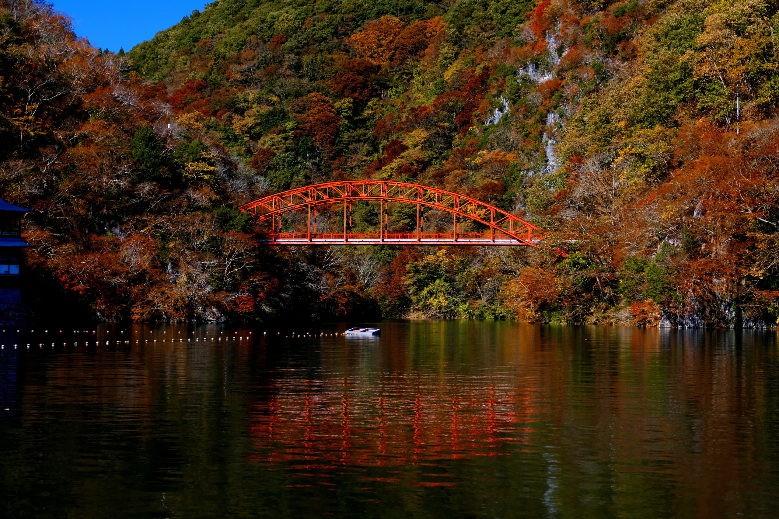 Red arched bridge across a calm lake, framed by vibrant autumn trees on a hillside with reflections in the water.