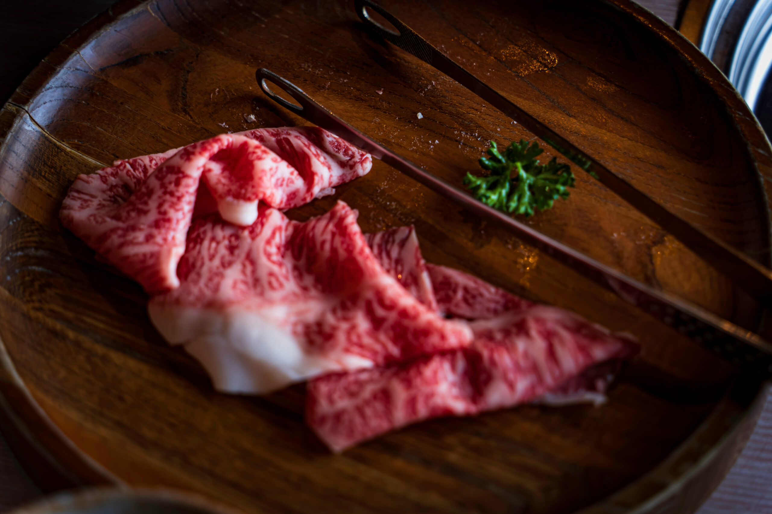 Raw marbled beef slices in a wooden bowl with tongs and a parsley sprig nearby.