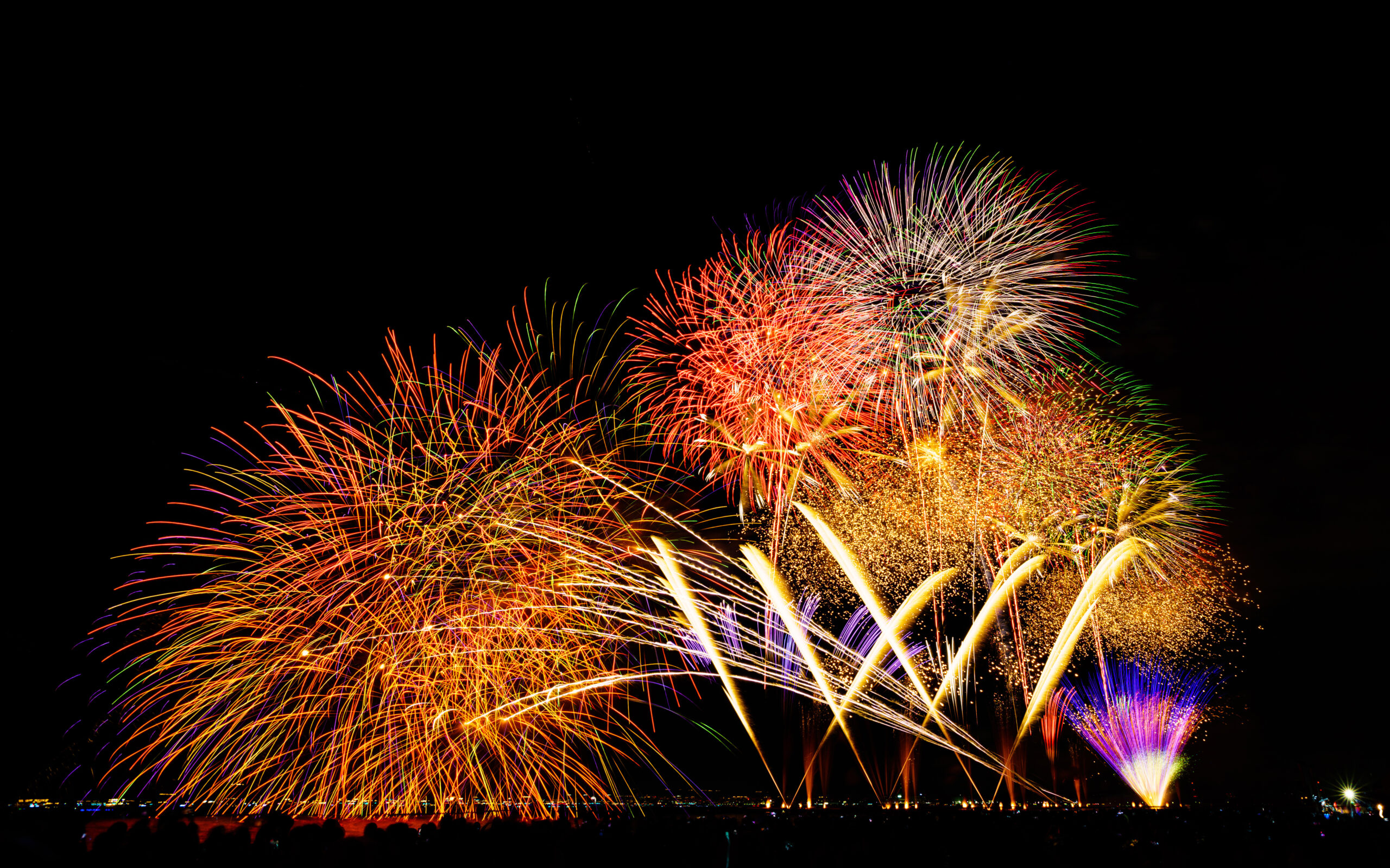 Colorful fireworks bursting over a dark night sky above a silhouetted crowd along a shoreline. Their reflections shimmer on the water below.