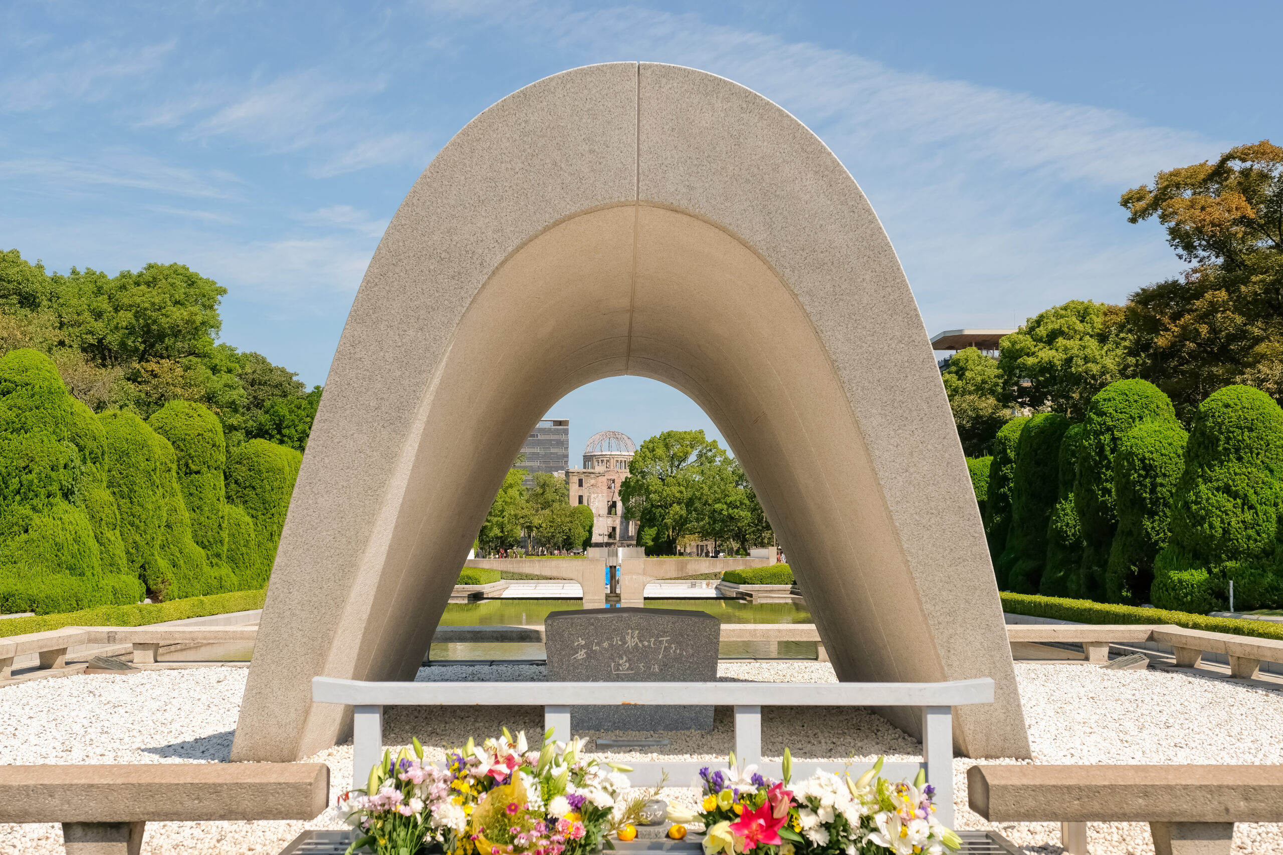 Stone arch sculpture over a shallow reflecting pool, with colorful flowers in the foreground and green trees on both sides in a memorial garden.