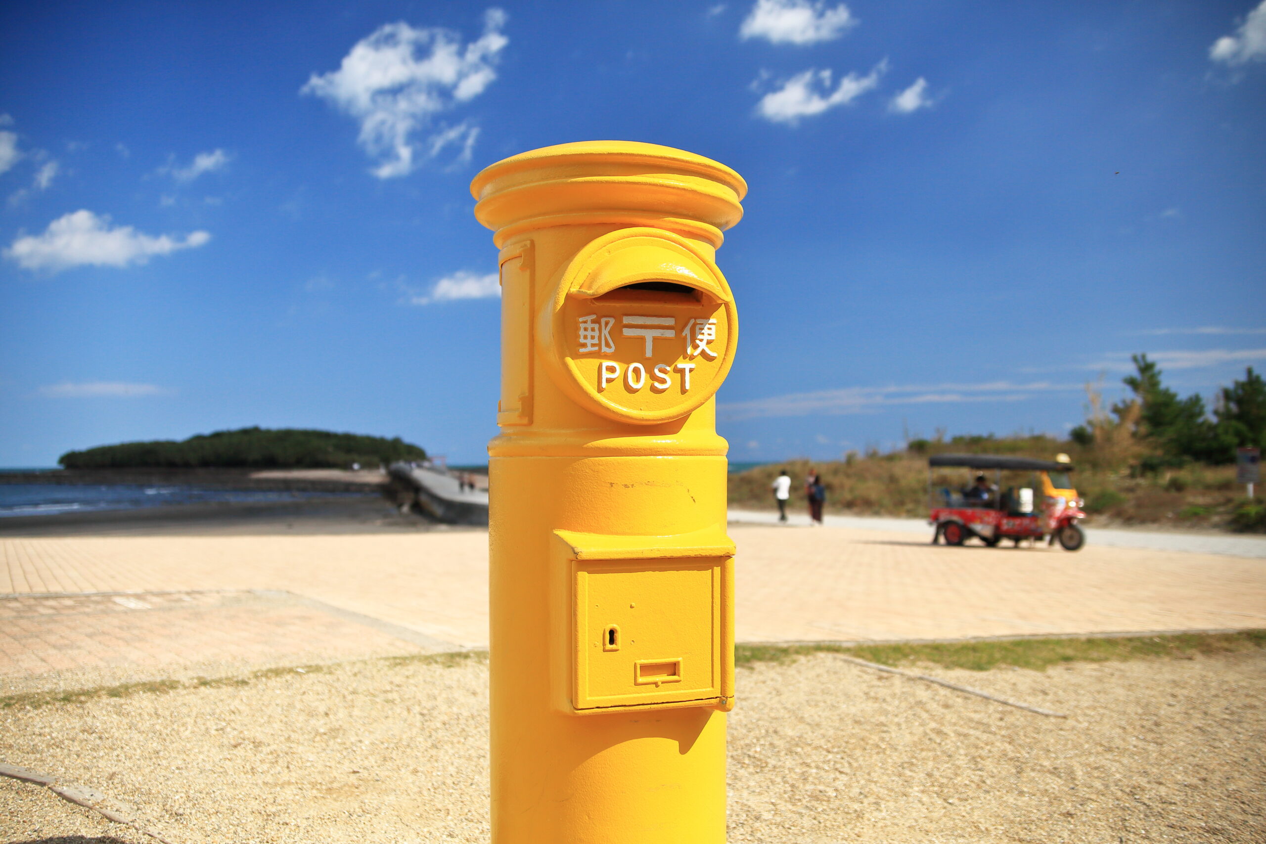 Yellow post box on a seaside promenade under a bright blue sky with scattered clouds.
