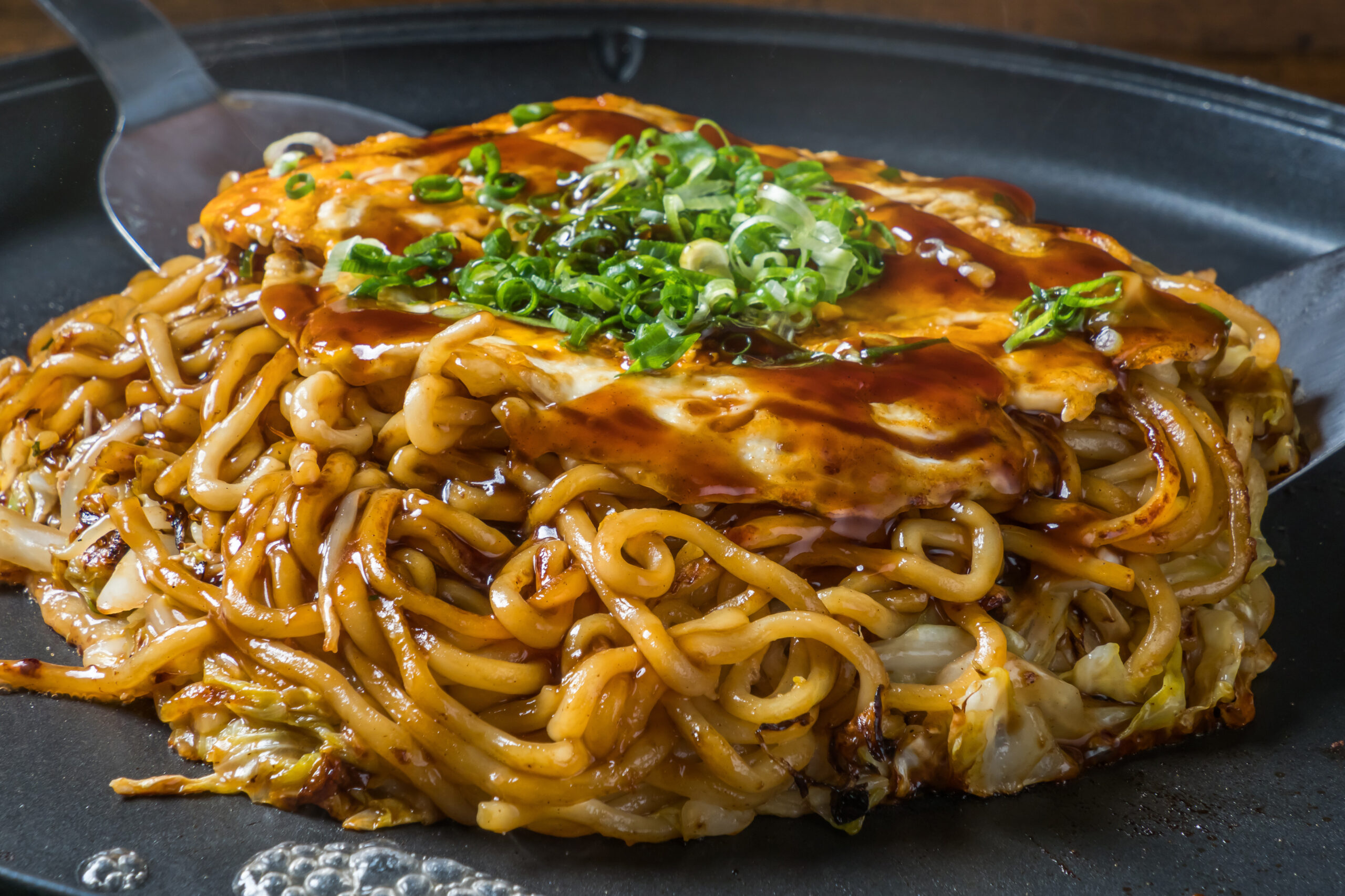 Stir-fried noodles in a dark sauce topped with chopped green onions on a black skillet