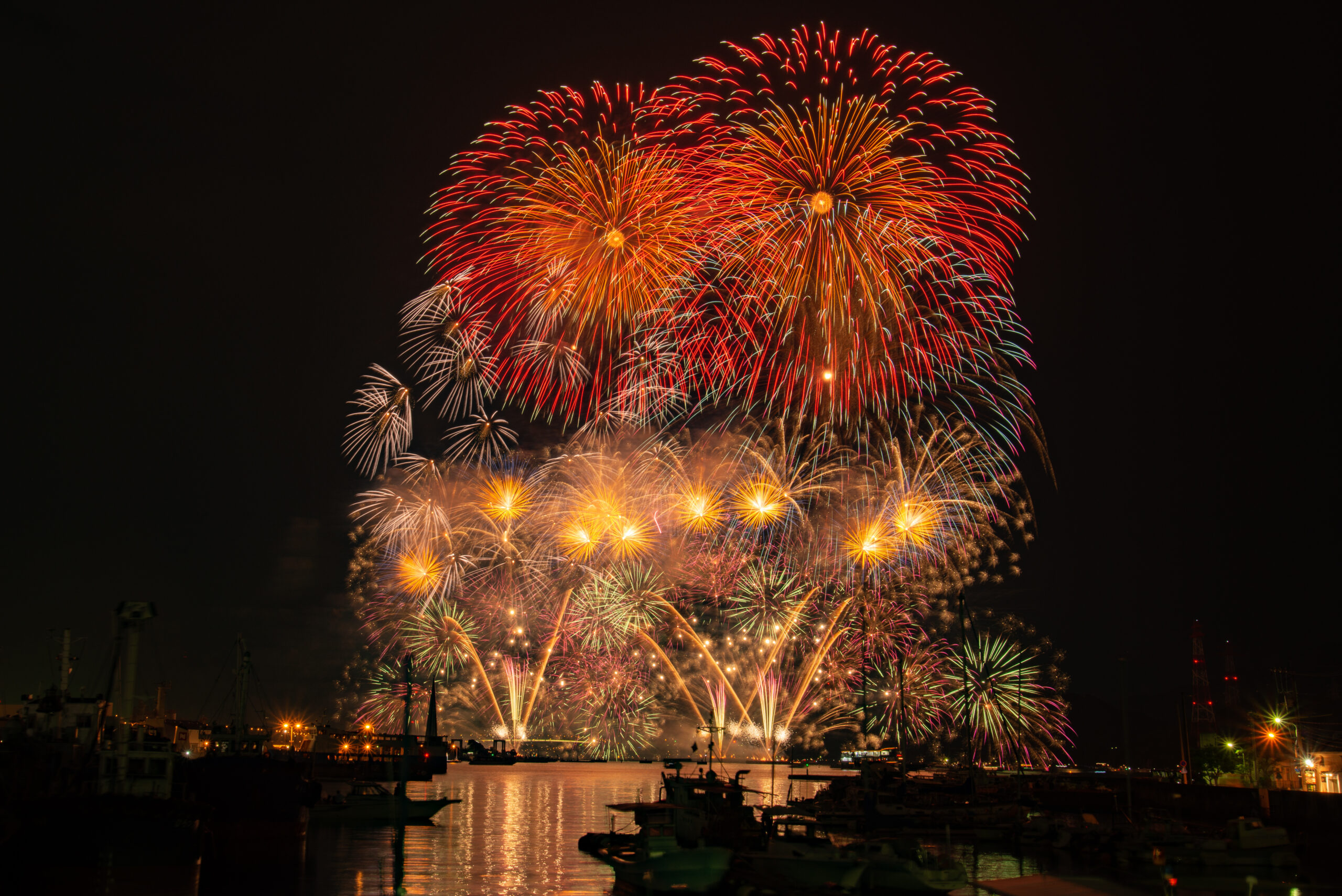 Vibrant fireworks over a dark harbor, reflecting on the water with boats anchored below at night.