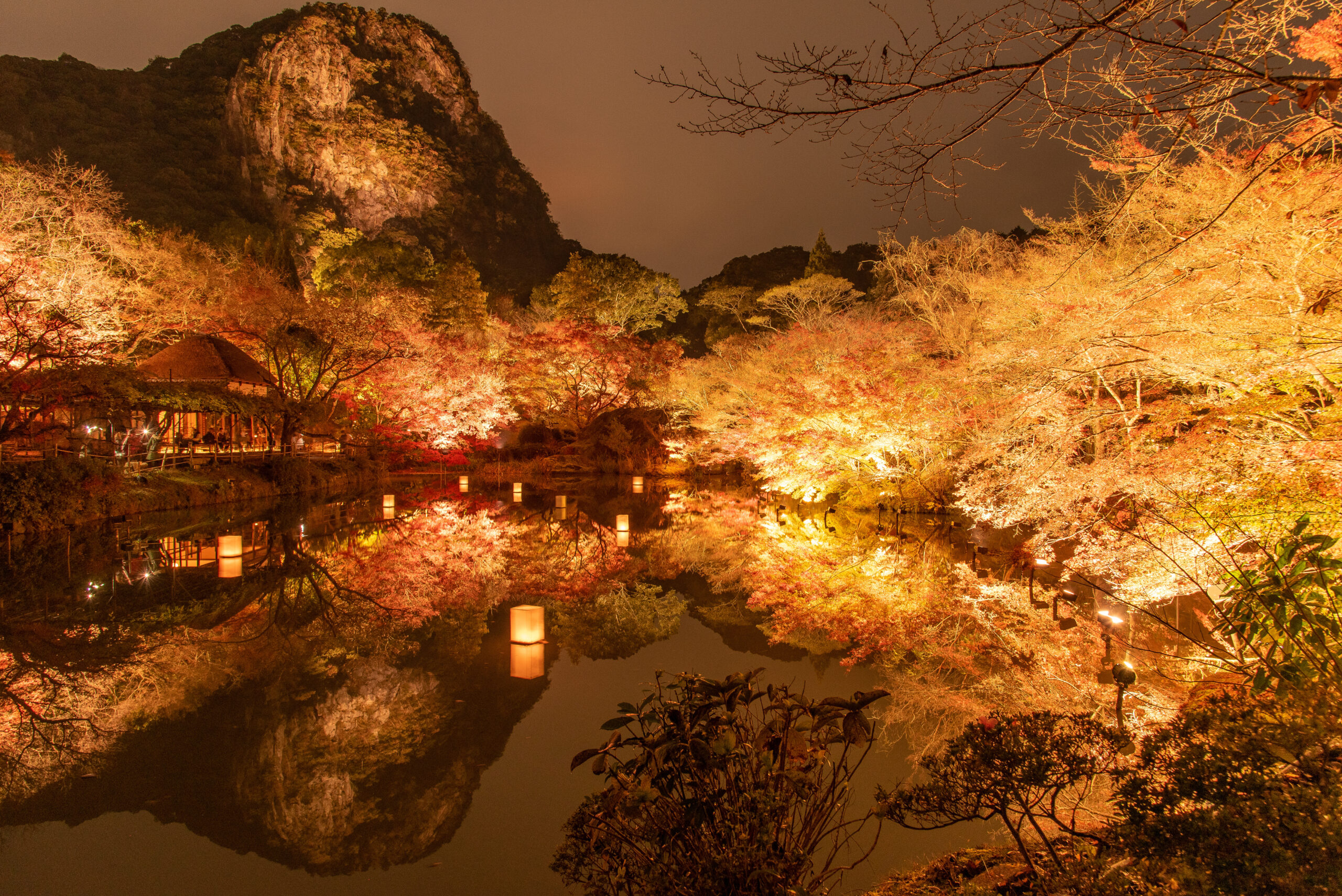 Night garden scene with illuminated orange and red autumn trees reflected in a calm pond, lanterns floating on the water, and a rocky hill in the background under a dark sky.