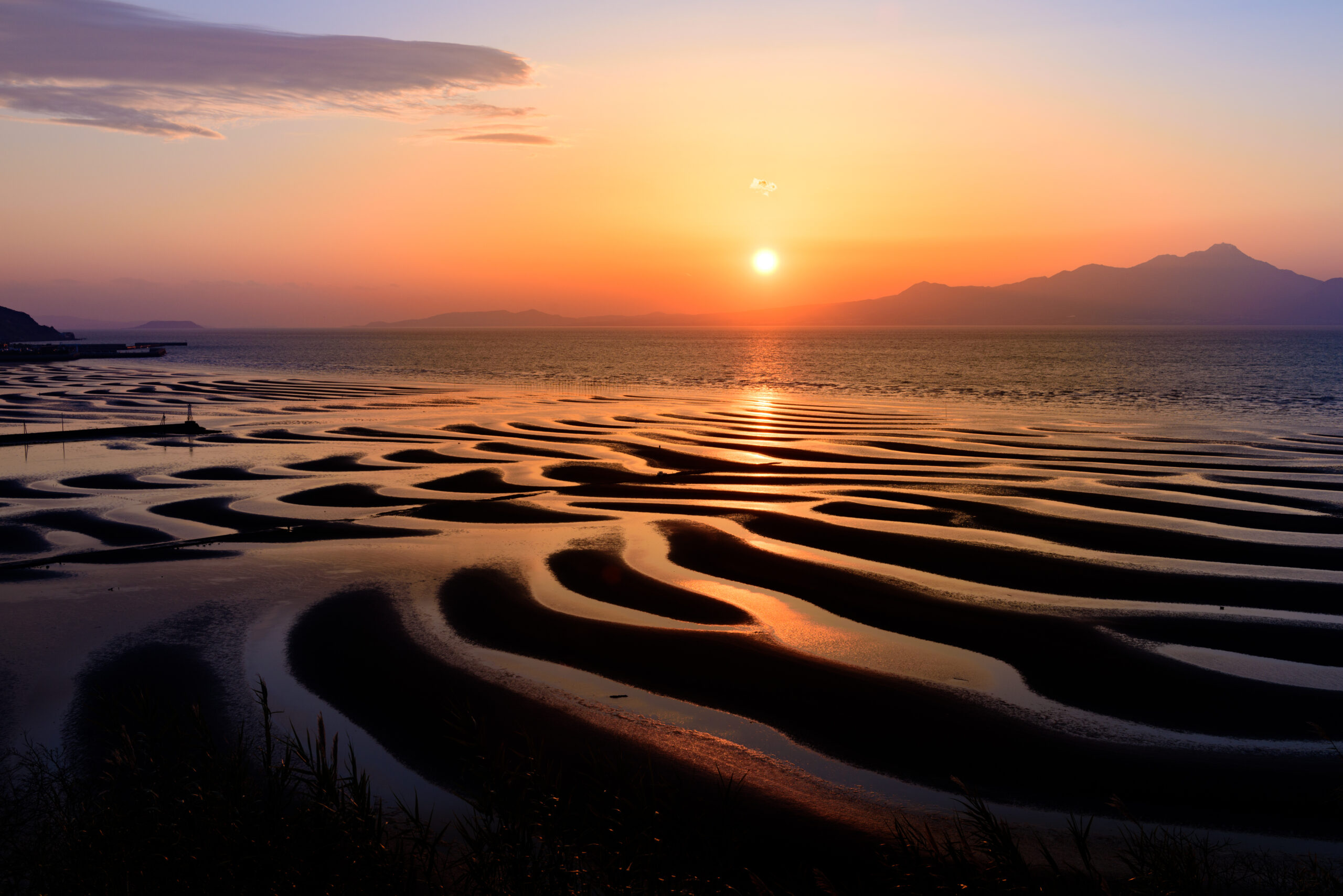 Sunset over the sea with orange glow, distant mountains, and tidal pools reflecting the sky in the foreground.