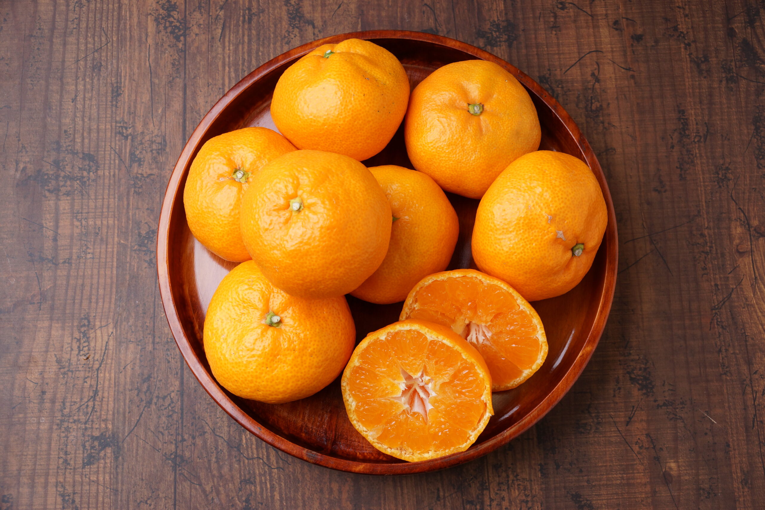 Brown bowl filled with bright oranges on a rustic wooden table, including a peeled orange showing its segments