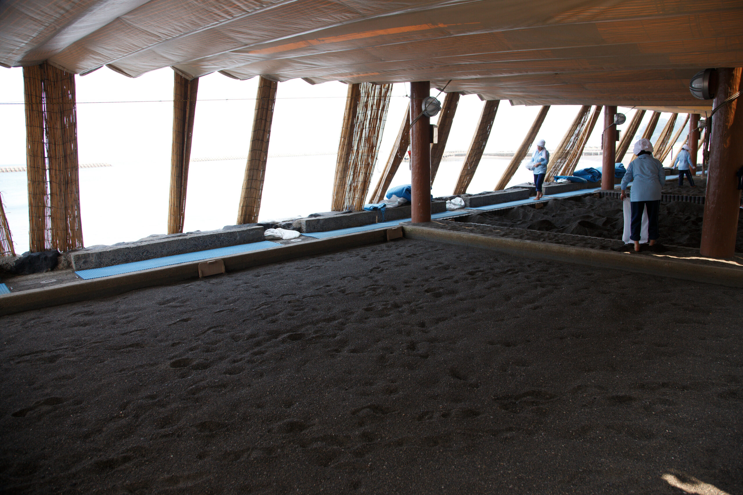 Staff in light blue uniforms and white hats walk along a wooden walkway under a thatched shelter at a beach.Workers inspecting a shaded area by the shore.