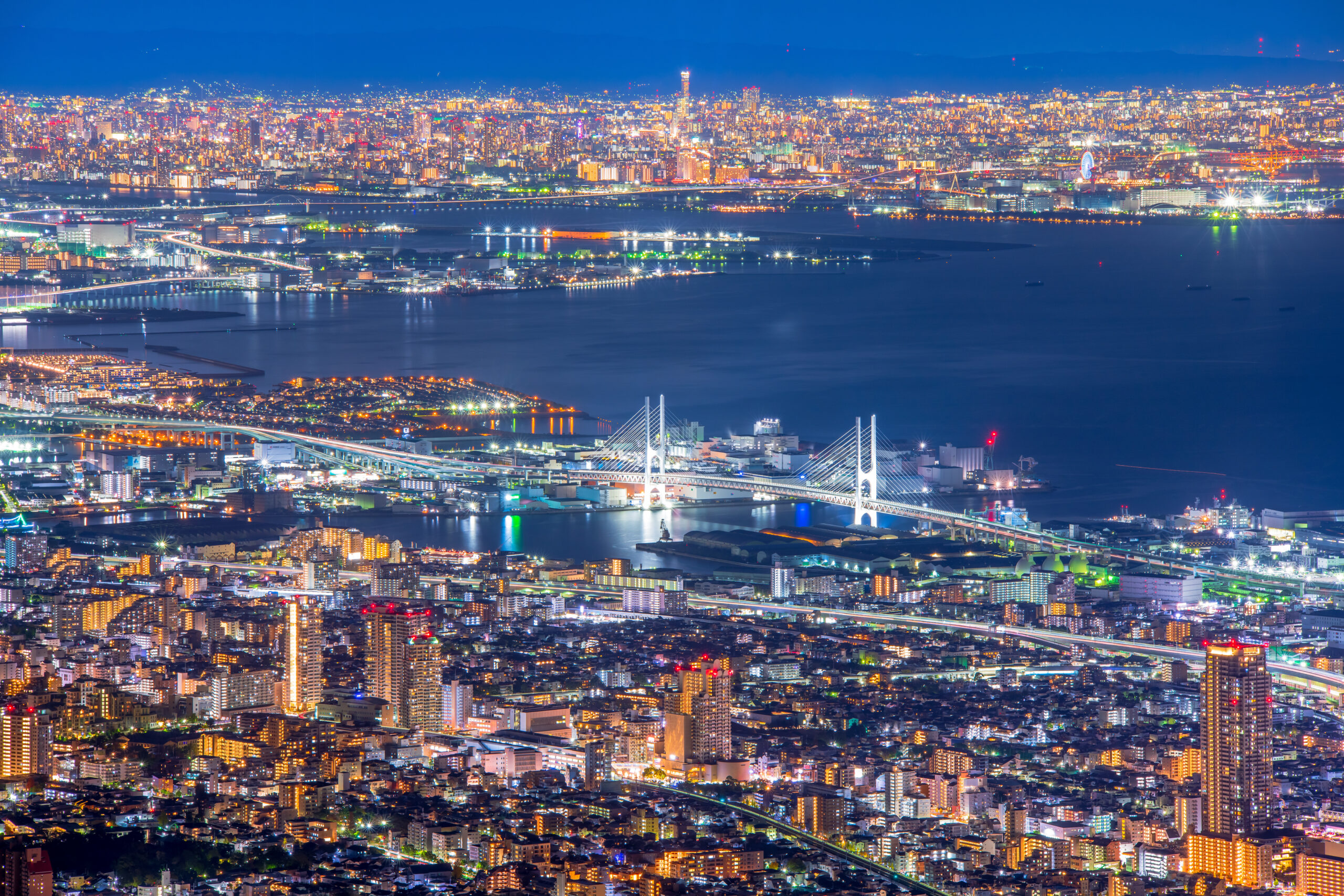 Aerial night view of a sprawling, brightly lit city with a large bridge crossing a bay and reflecting lights on the water.