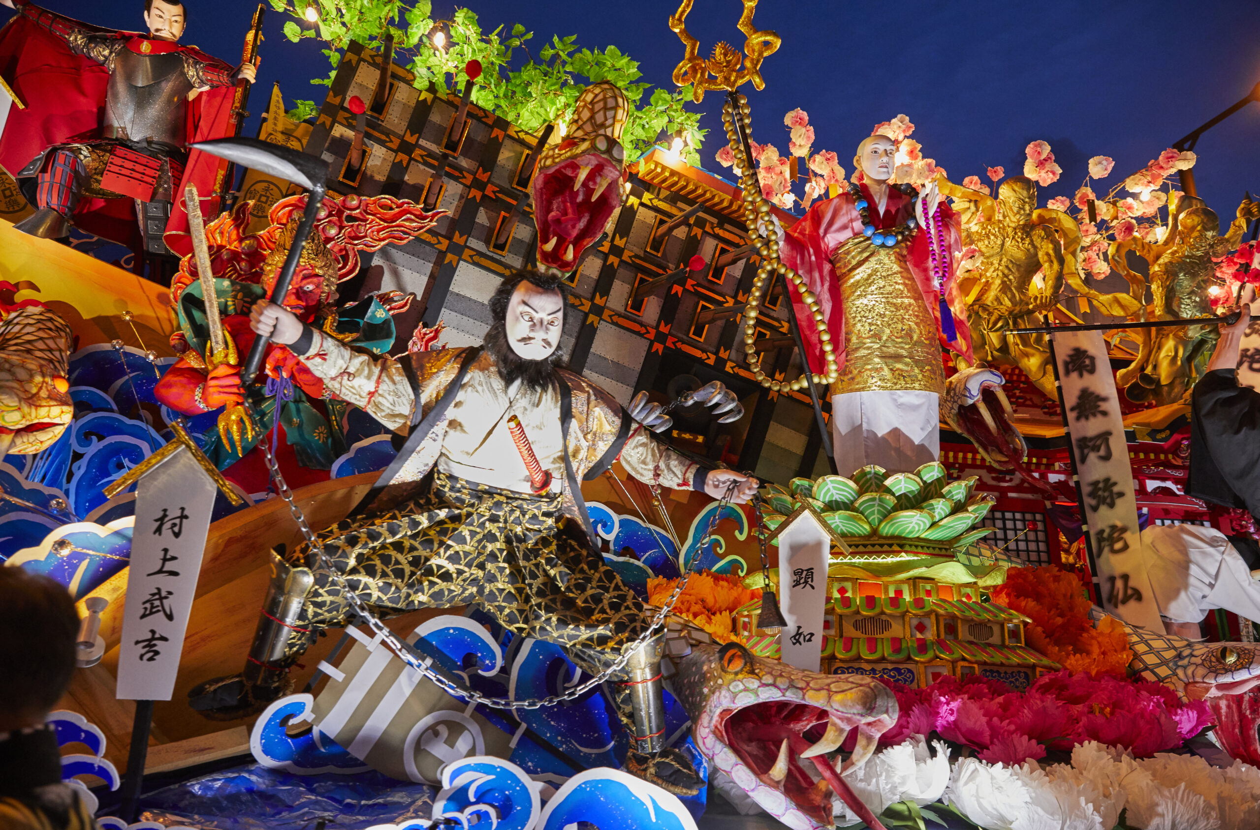 Elaborate festival float with masked performers, dragons, and gold statues lit at night in a parade.