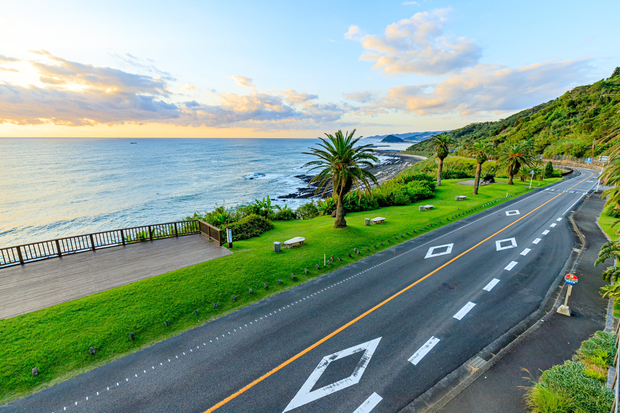A coastal road runs along a grassy park with palm trees on the right, the ocean to the left and hills in the distance under a partly cloudy sky at sunset/sunrise.
