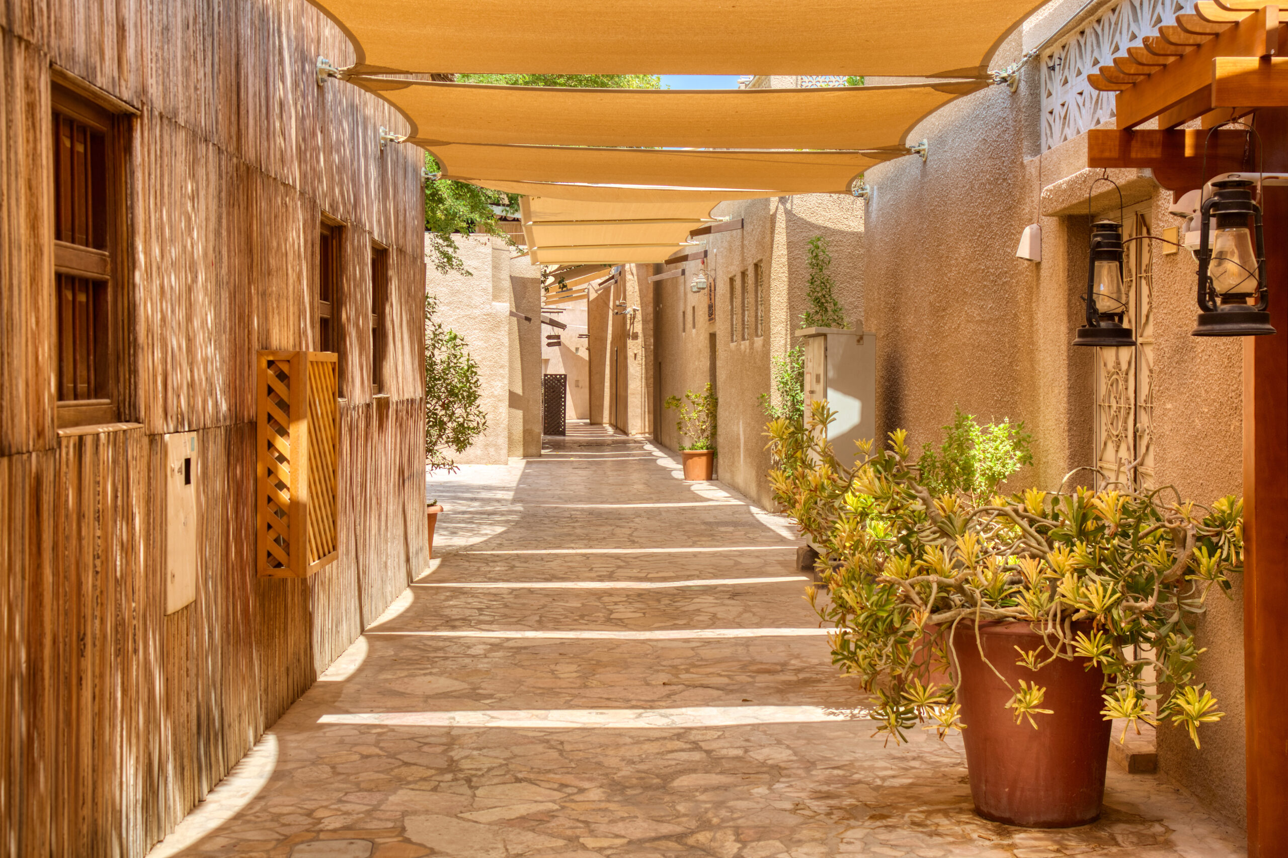 Narrow sunlit alley with wooden left wall, beige stucco right wall, beige fabric shade canopies overhead, and potted plants along the walkway.