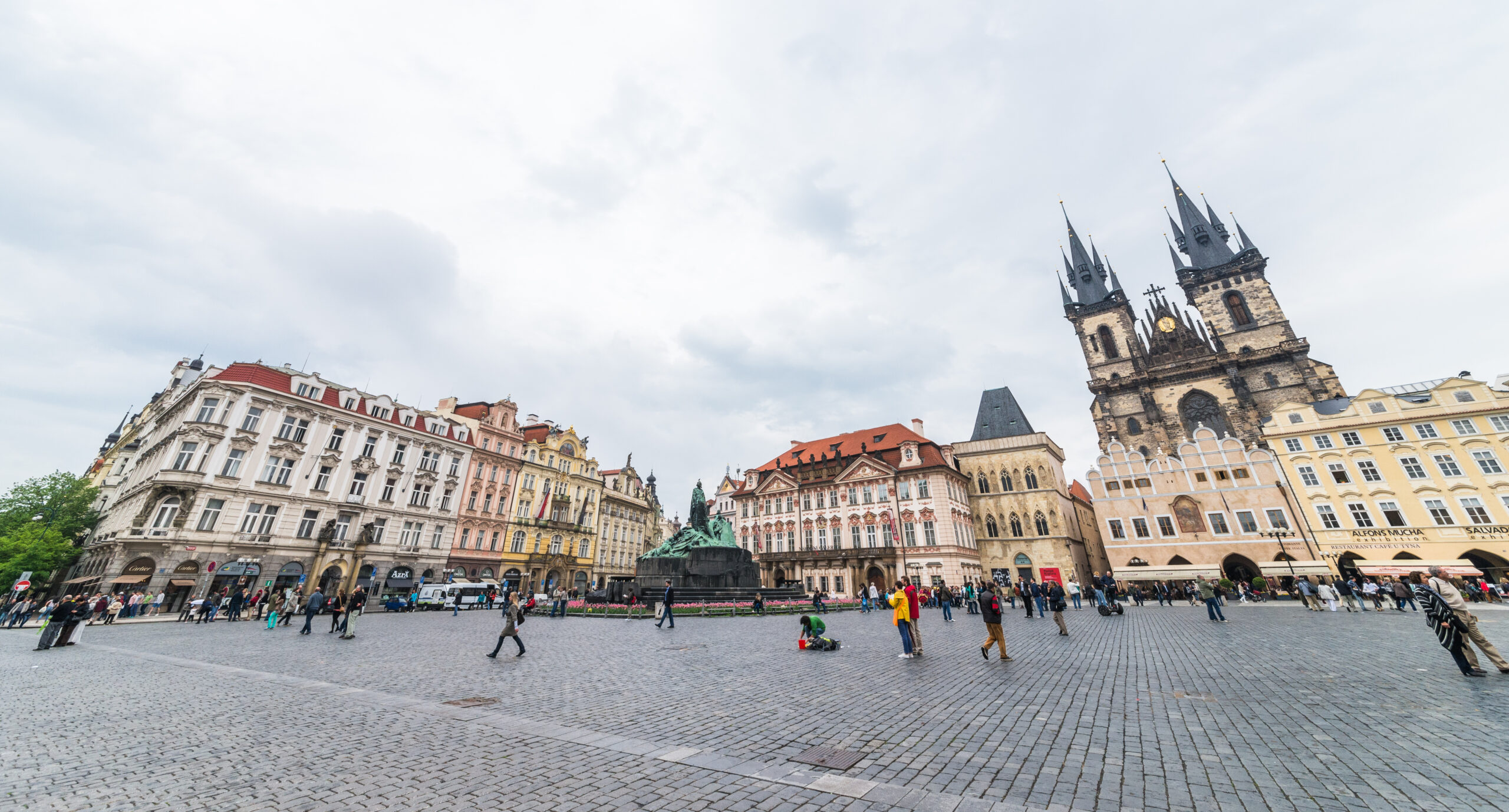 Colorful historic buildings surround a wide cobblestone square, with a central statue and a Gothic church with twin spires; people walk about under a cloudy sky.