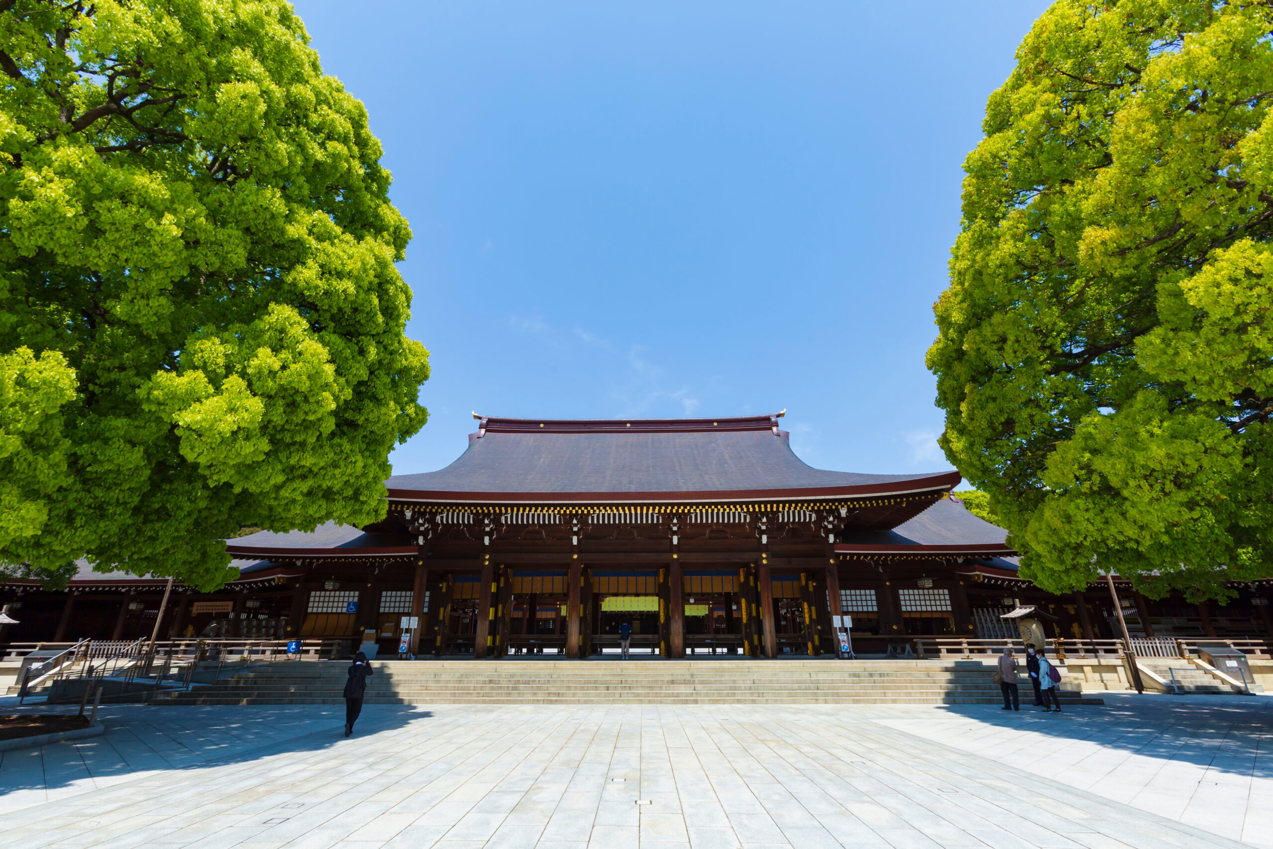 Front view of a traditional Japanese temple with red wooden architecture, flanked by bright green trees and a blue sky.