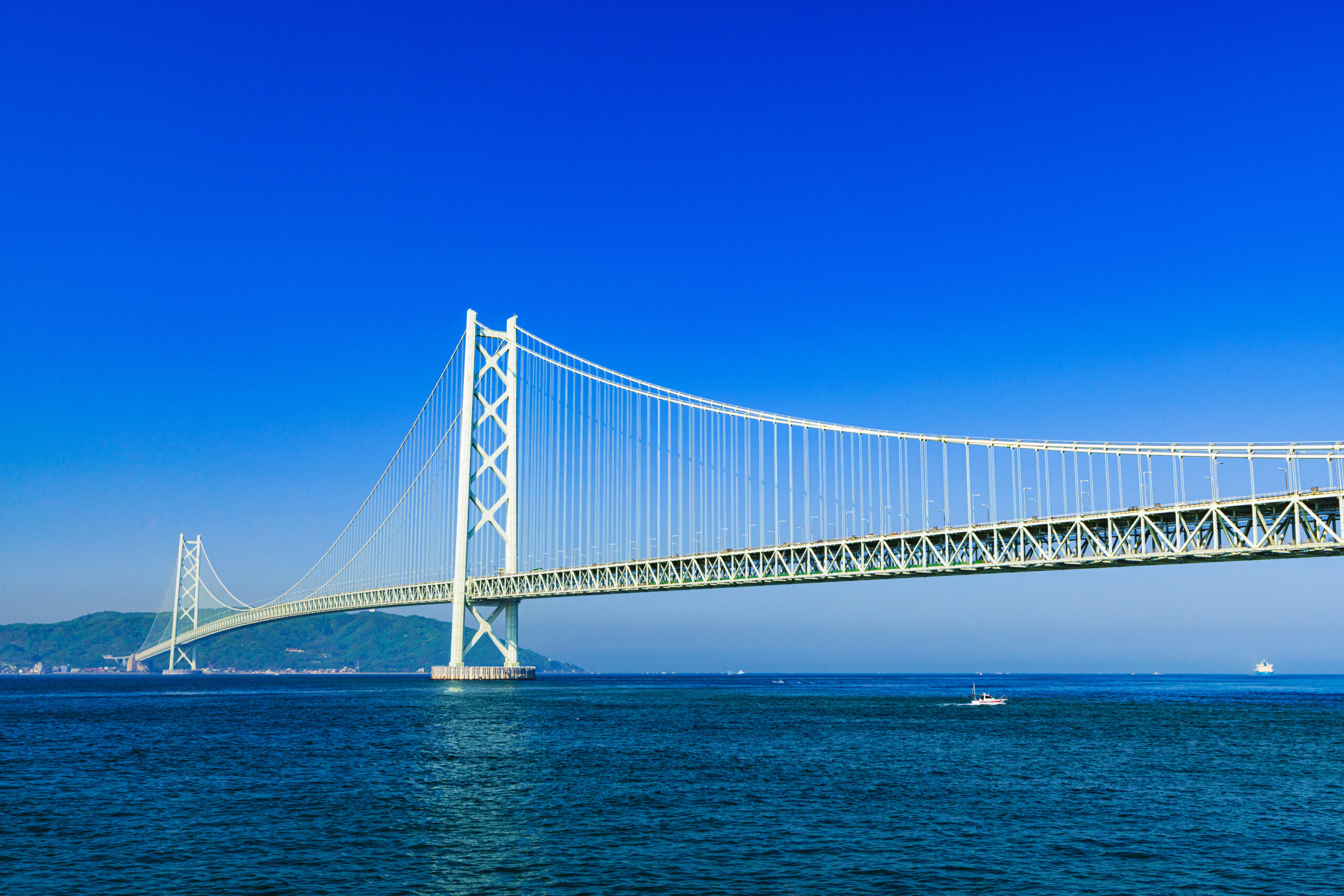 White suspension bridge spanning a calm blue sea under a clear blue sky, with hills in the distance and a small boat nearby.